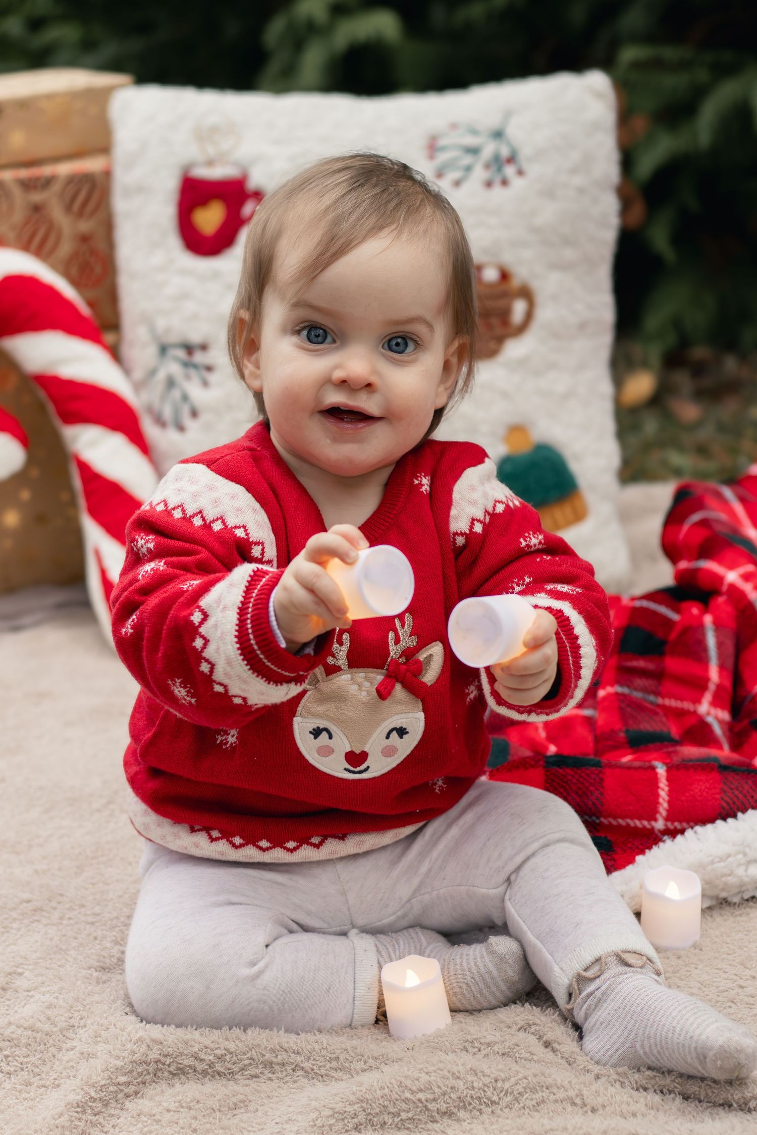 An adorable baby in a red reindeer sweater plays with candles among Christmas decorations.