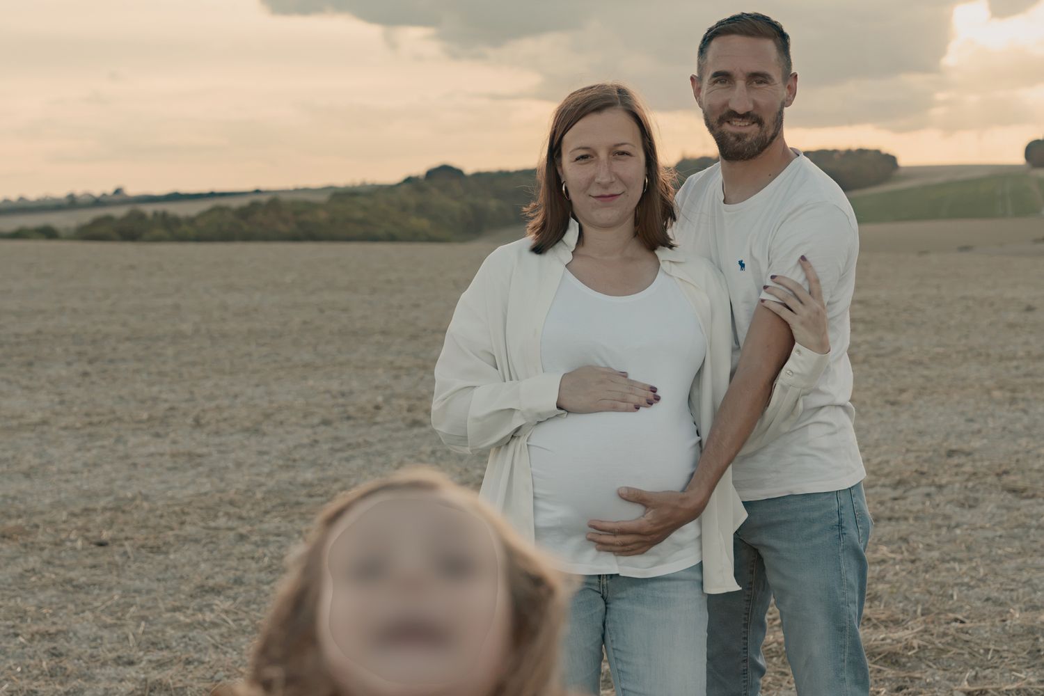 Pregnant couple standing in a field at sunset, with a blurred child in the foreground.
