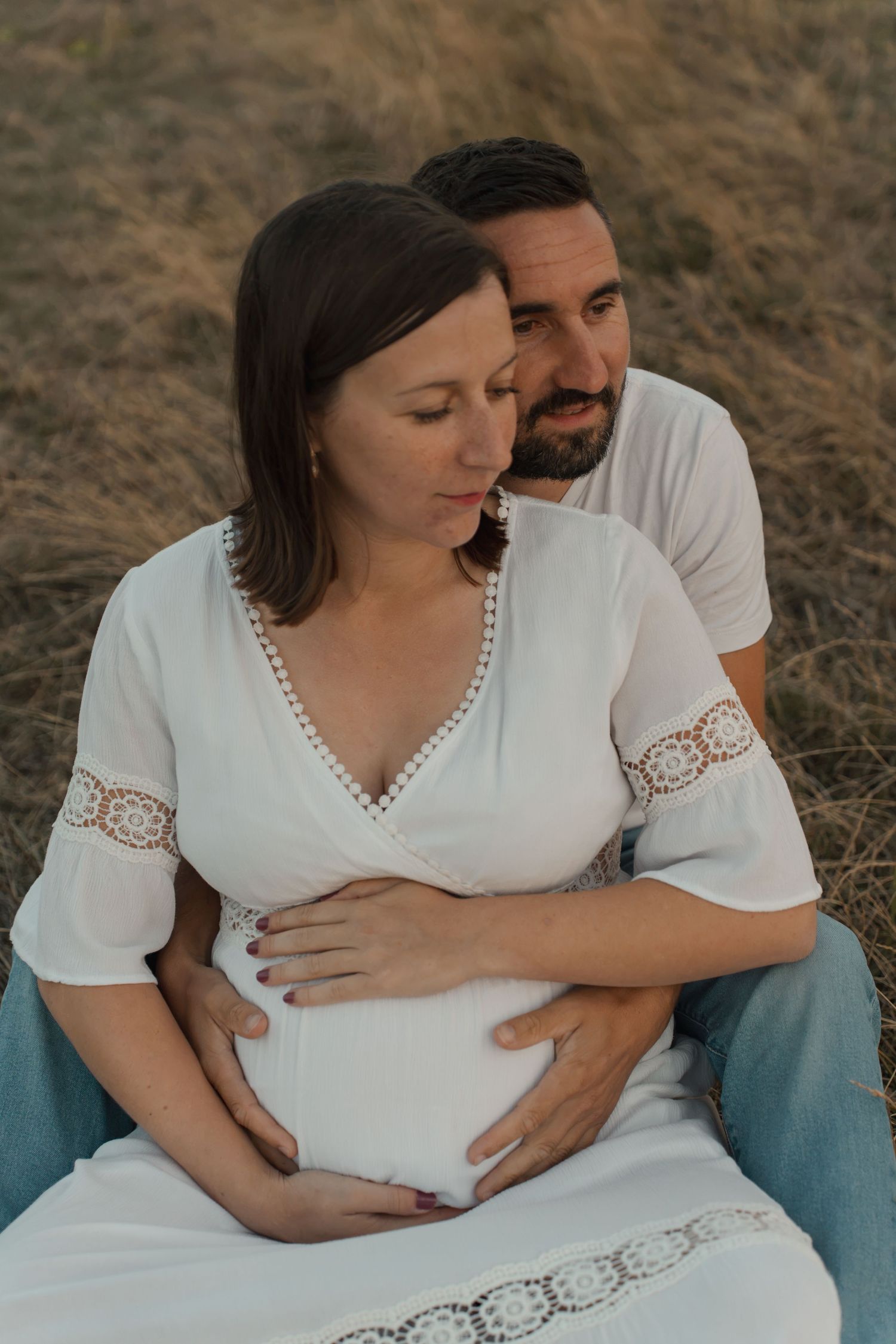 Couple sitting in field, man embracing pregnant woman in white dress, both holding her baby bump.