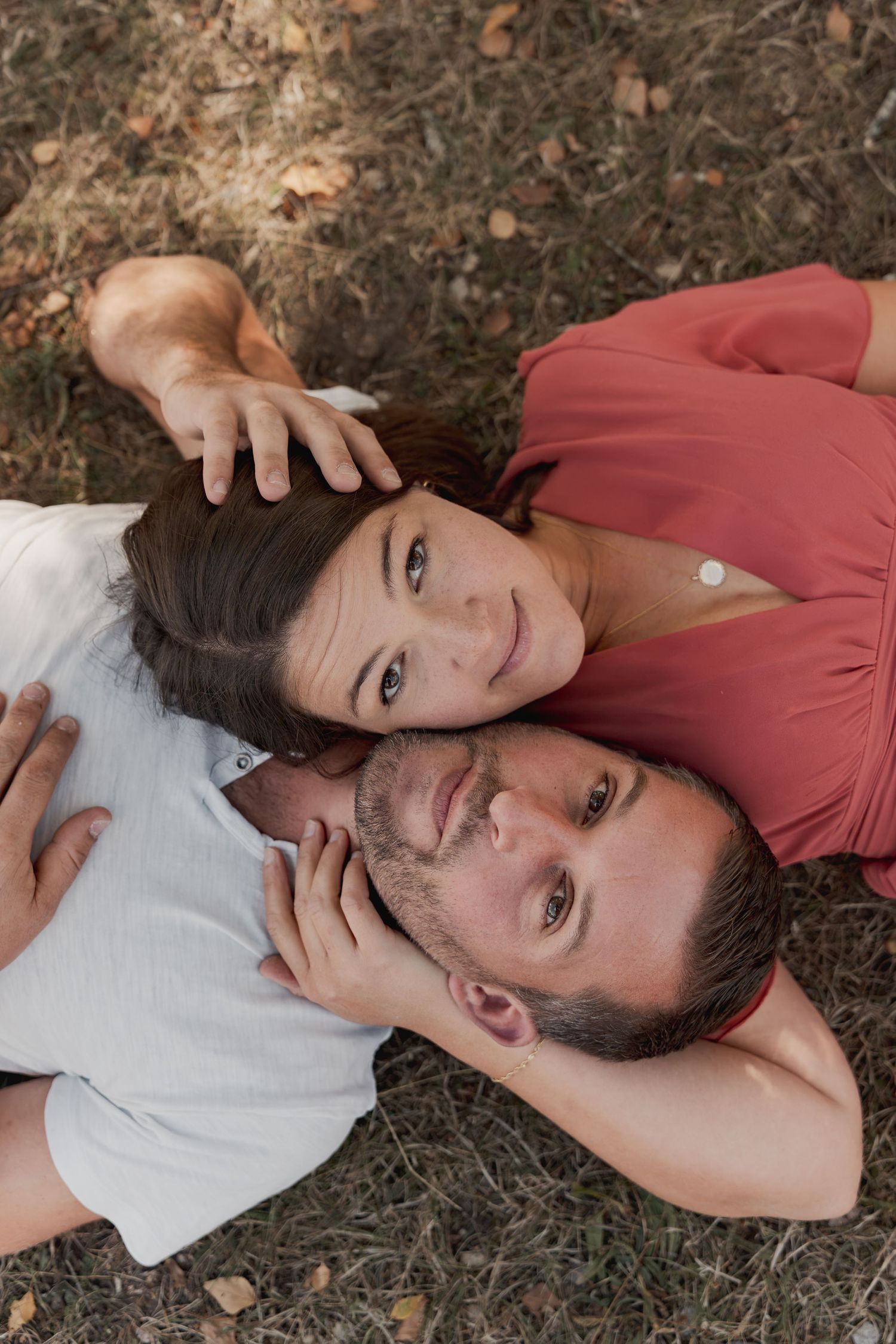 Couple lying on grass outdoors, woman in red top resting head on man in white shirt, both smiling up at camera.