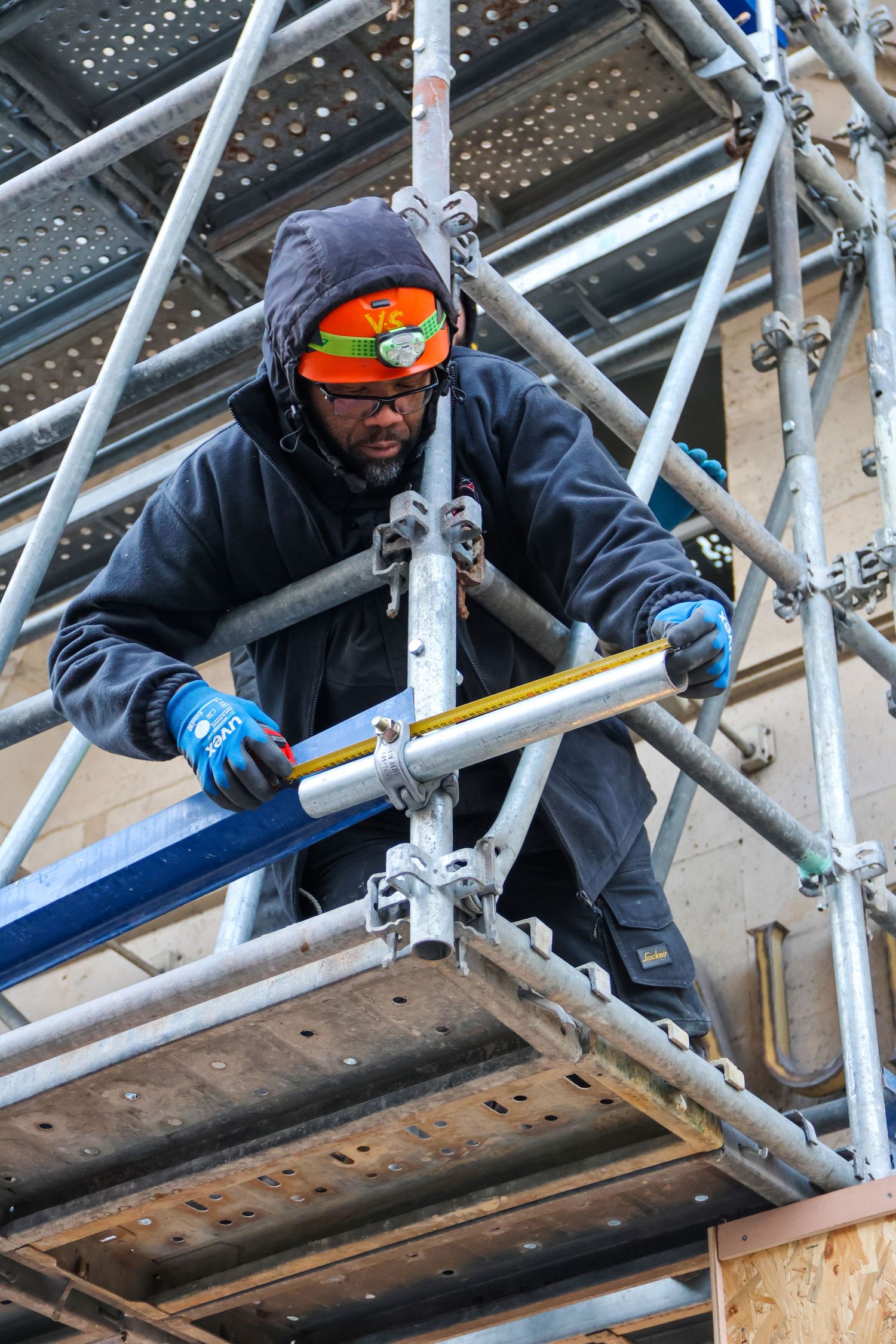 Construction worker in safety gear assembling scaffolding on a building site, wearing orange goggles and hard hat.