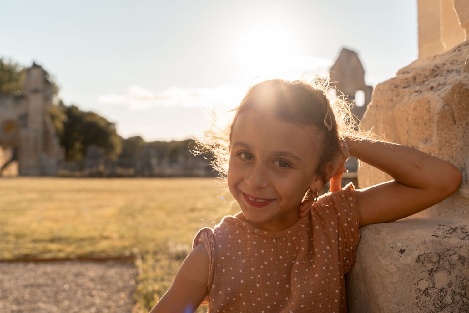 A smiling young girl with pigtails leans against a stone wall in golden sunlight outdoors.