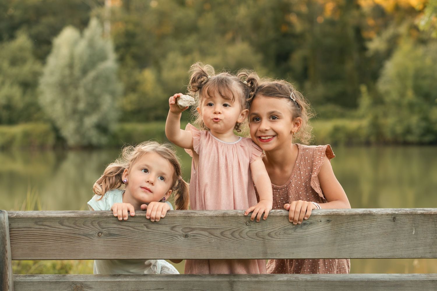 Three young girls lean over a wooden fence near a lake, making playful expressions in a lush outdoor setting.