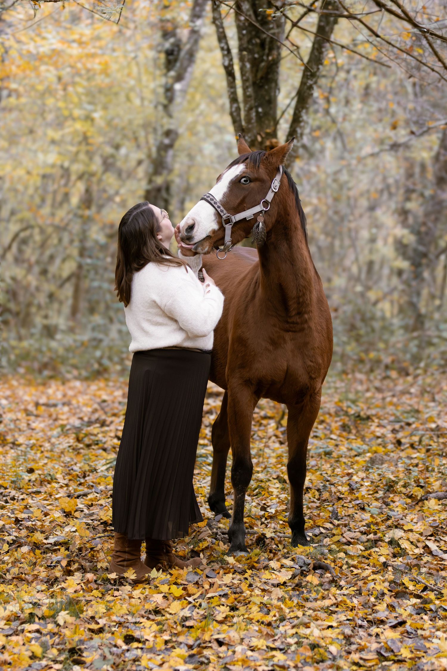 Woman in cream sweater and dark skirt standing with a chestnut horse in fall forest.