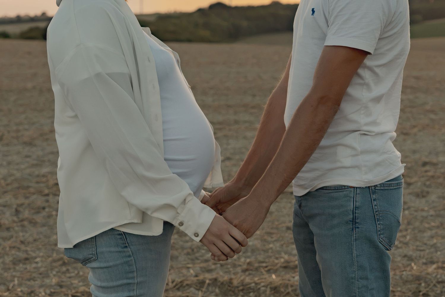 Pregnant couple holding hands in a golden field at sunset, wearing white shirts and jeans.