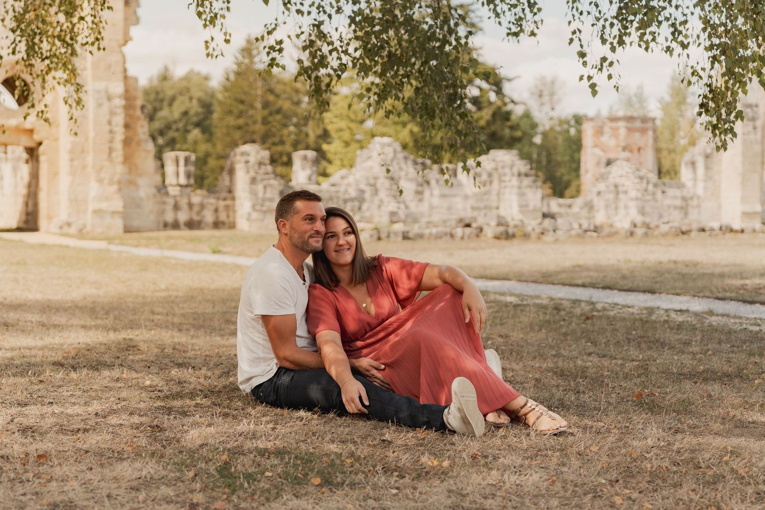 A couple sits together on grass near ancient stone ruins, smiling warmly on a sunny day.