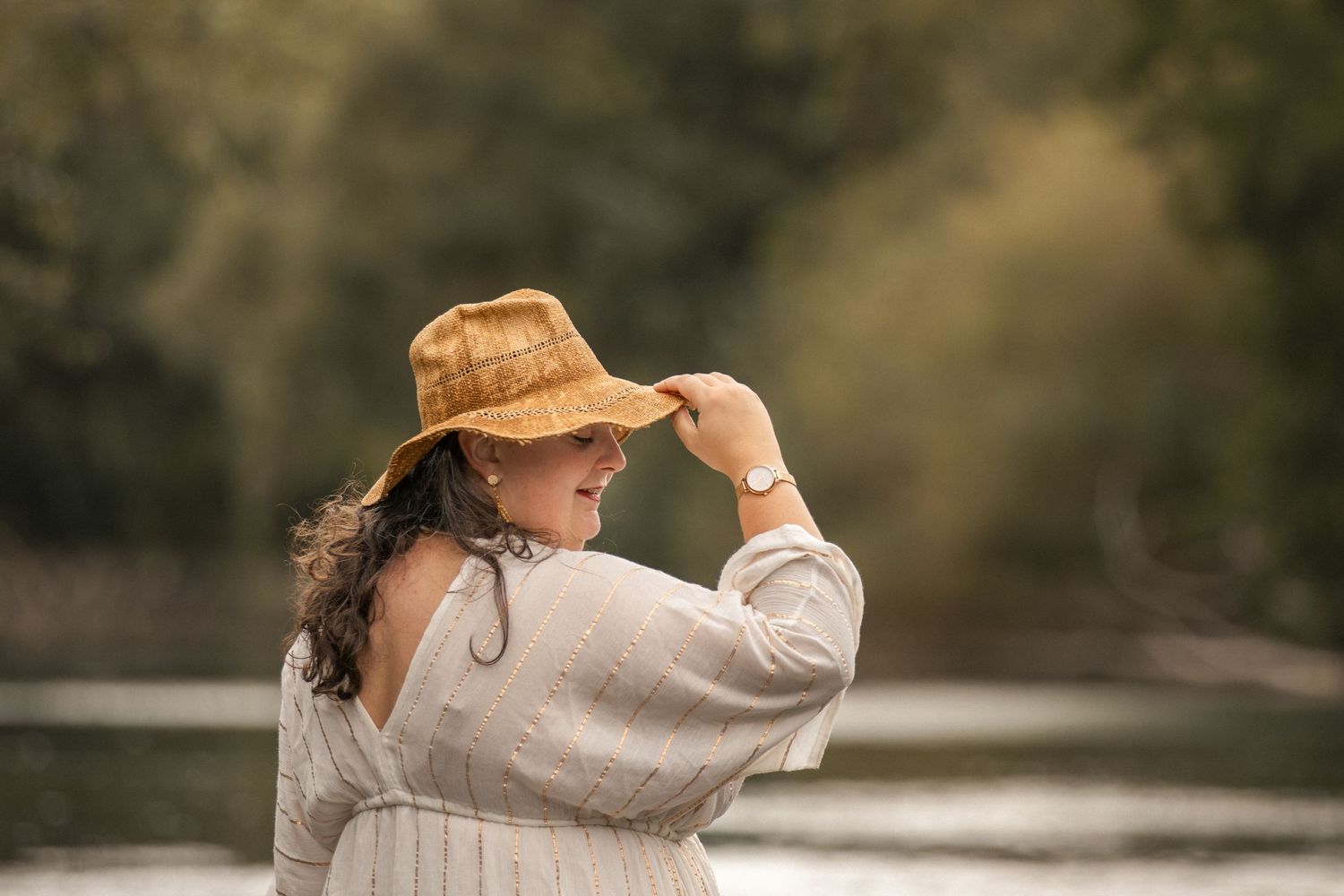 Woman in white dress and tan hat standing by a river, looking over her shoulder in a serene outdoor setting.