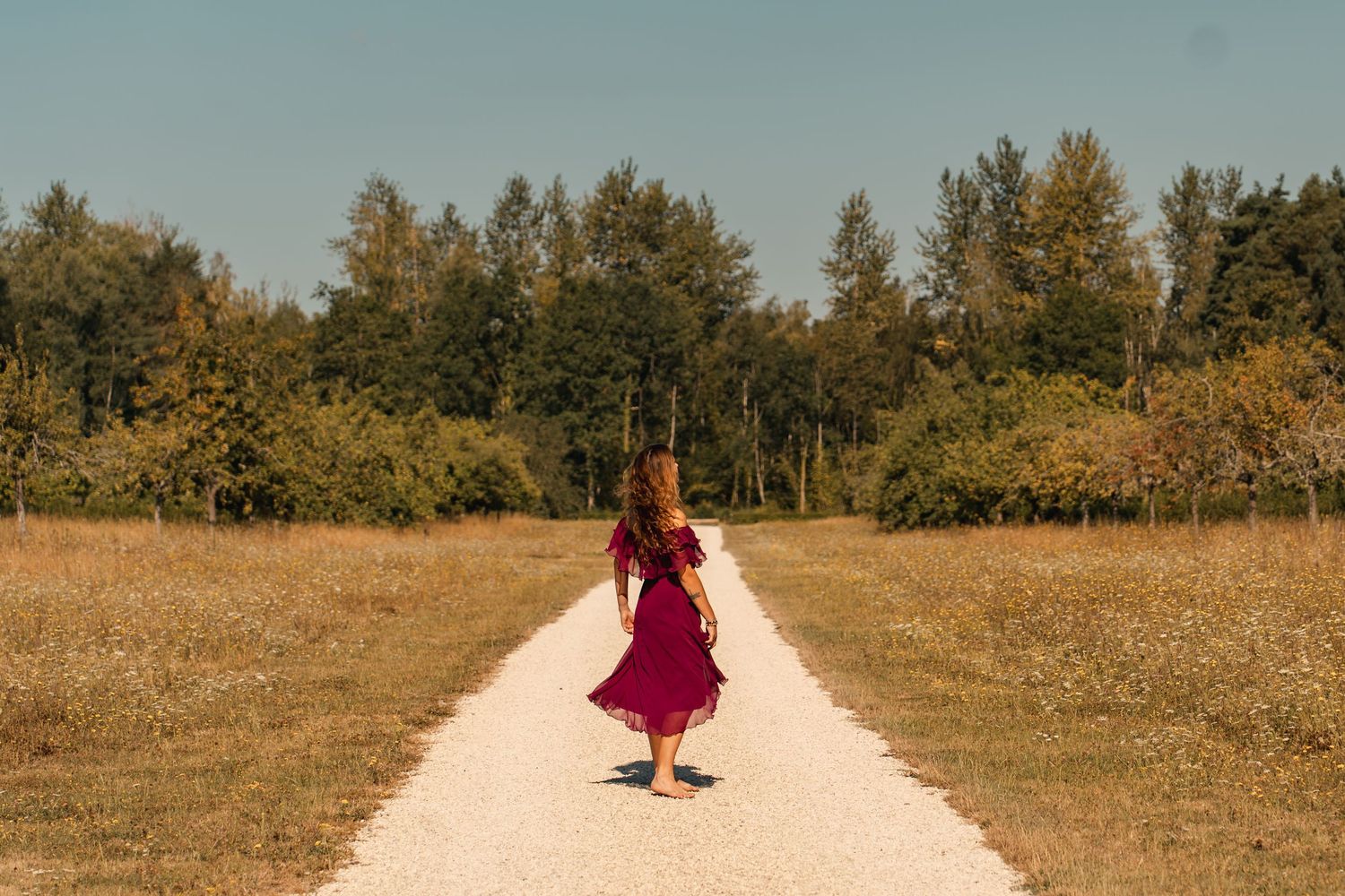Woman in red dress walking barefoot on a gravel path surrounded by lush green trees and golden fields.