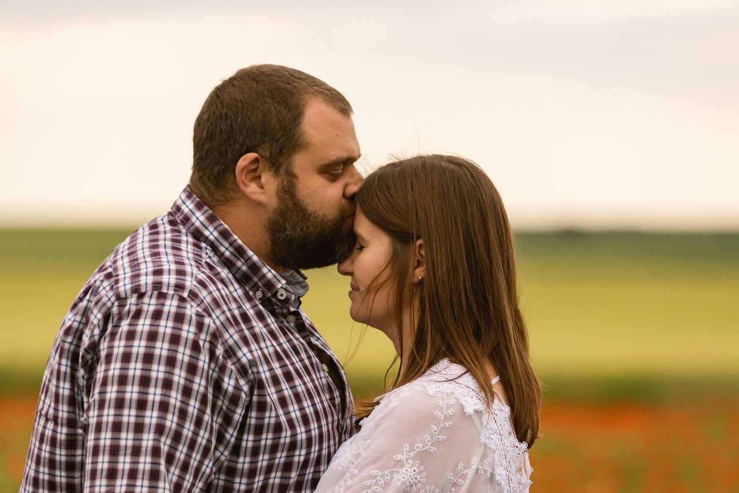 Bearded man in plaid shirt leaning in to kiss a woman in a white lace dress in a green field.