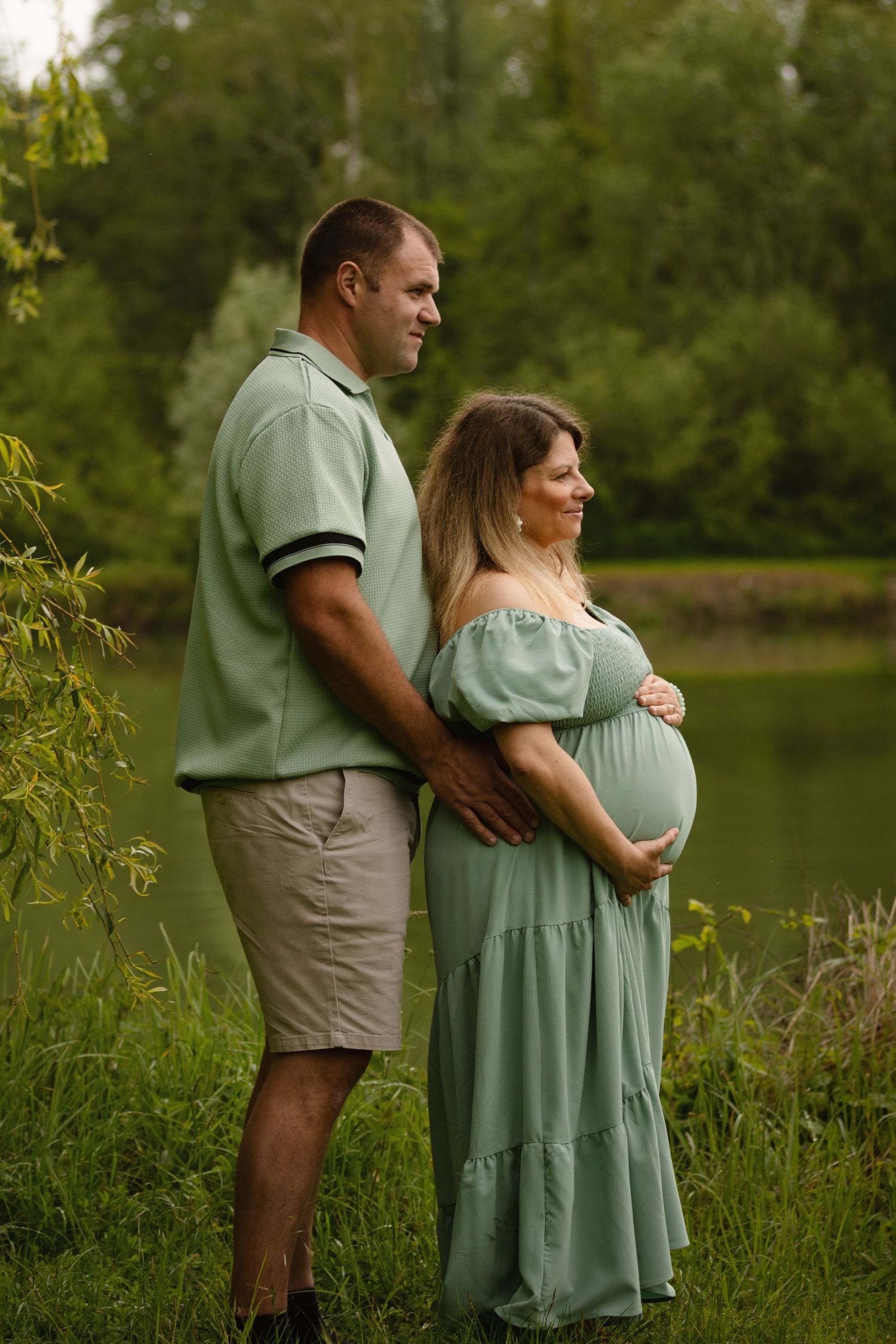 Couple in matching sage green outfits posing outdoors near a pond, woman in off-shoulder maternity dress cradling baby bump.
