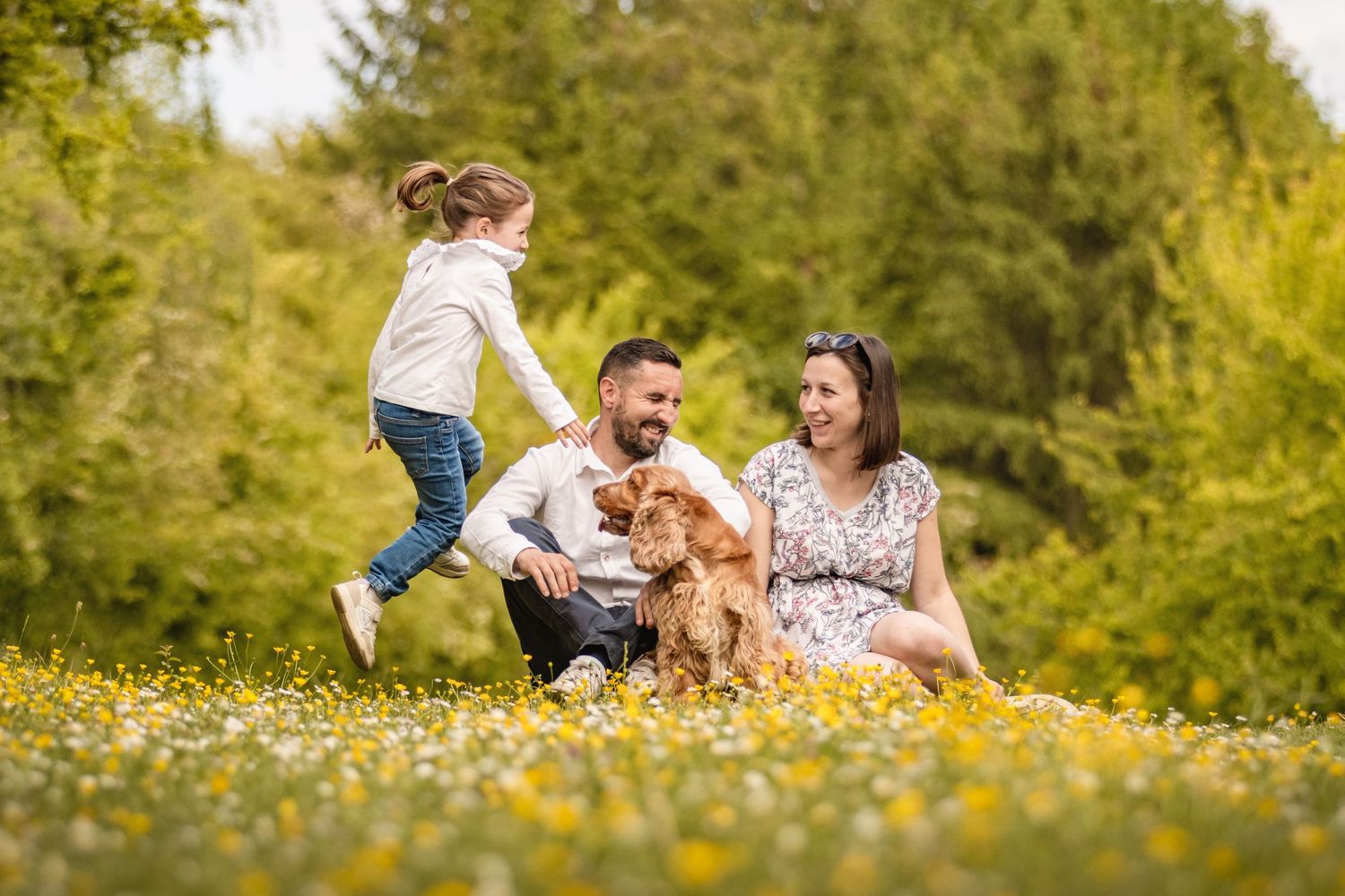 Family with a young girl and golden dog enjoying a sunny day in a flower meadow surrounded by green trees.