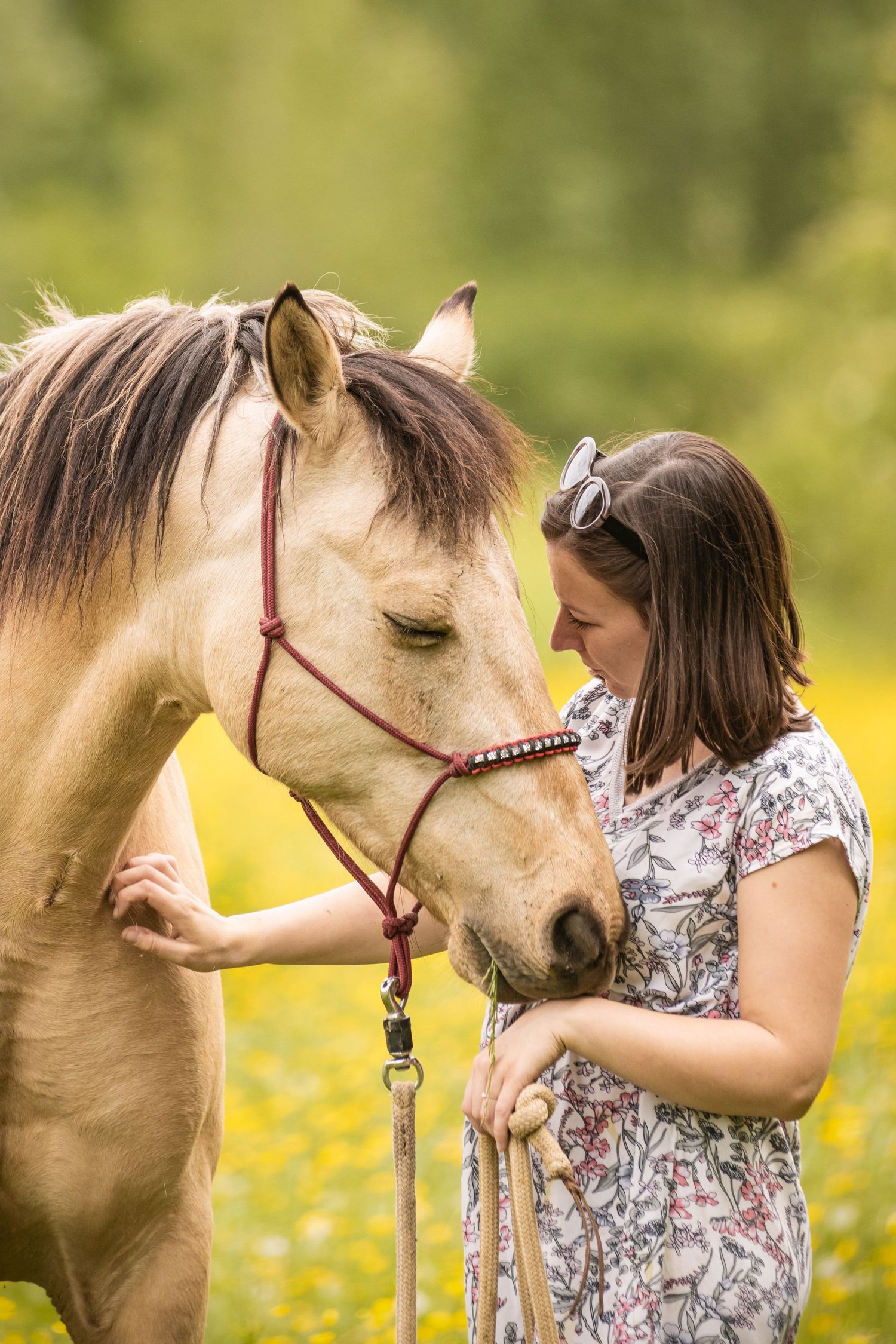 Young woman in floral dress tenderly bonding with a palomino horse in a sunny green meadow.