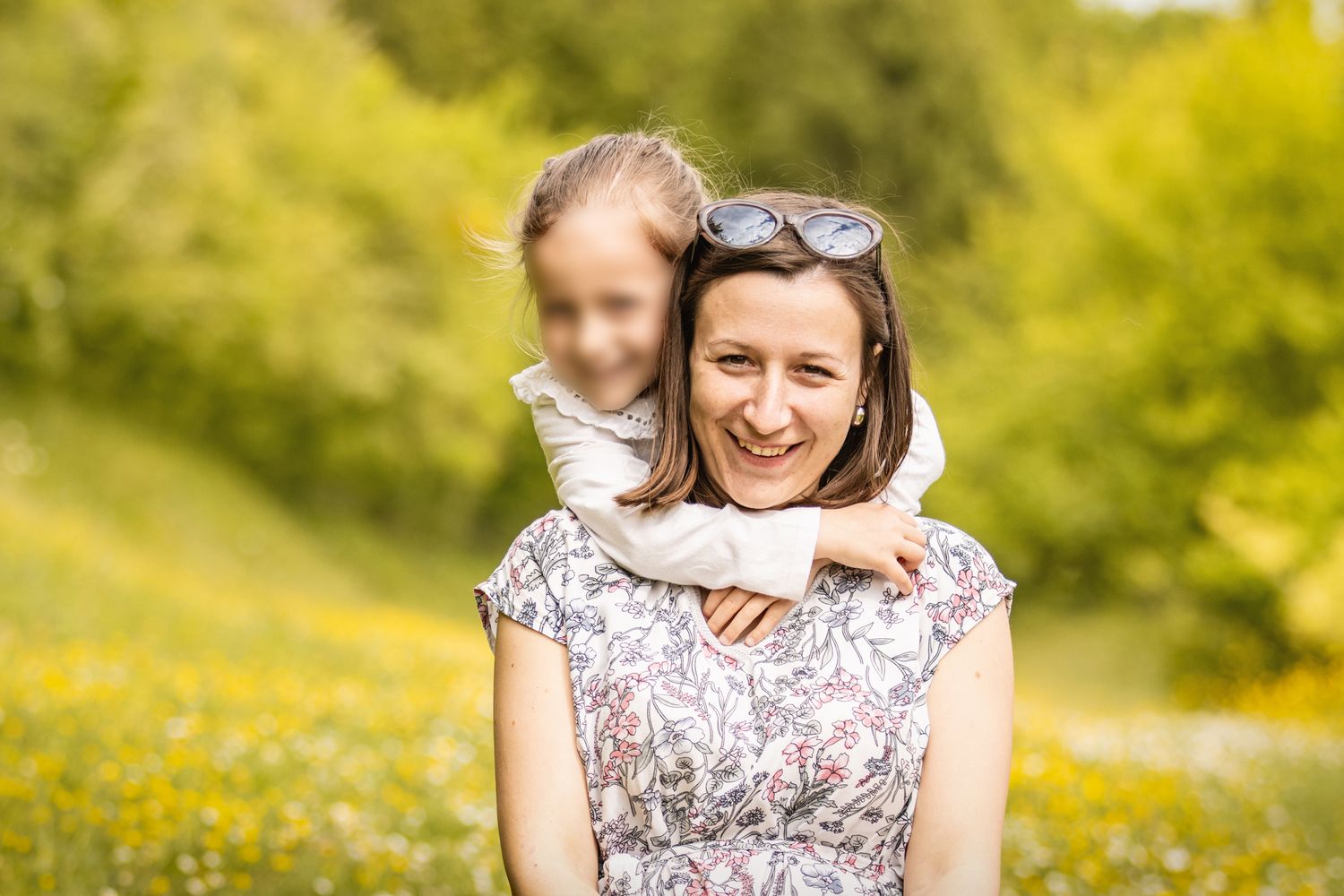 A smiling woman in a floral dress carries a young girl on her back in a sunny green meadow with yellow wildflowers.
