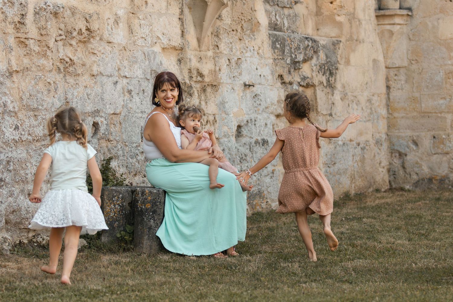 A mother in a mint dress sits outdoors holding a baby while two young girls in dresses play around her near a stone wall.