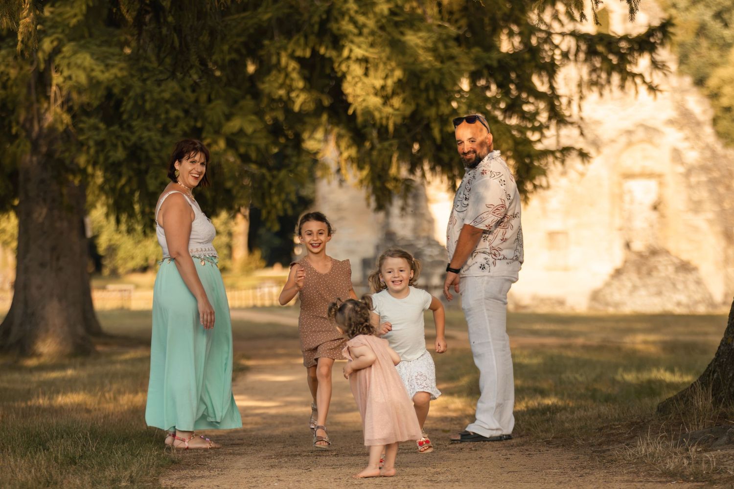 Family of four enjoying a sunny outdoor photoshoot among trees, with children playing and parents smiling warmly.