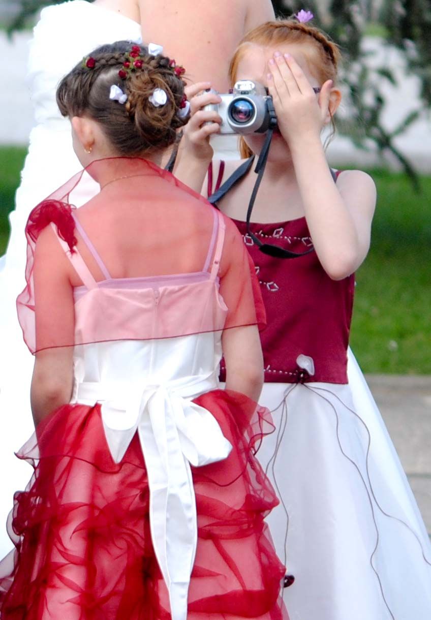 Two young flower girls in red and white dresses, one photographing with a camera at a wedding.