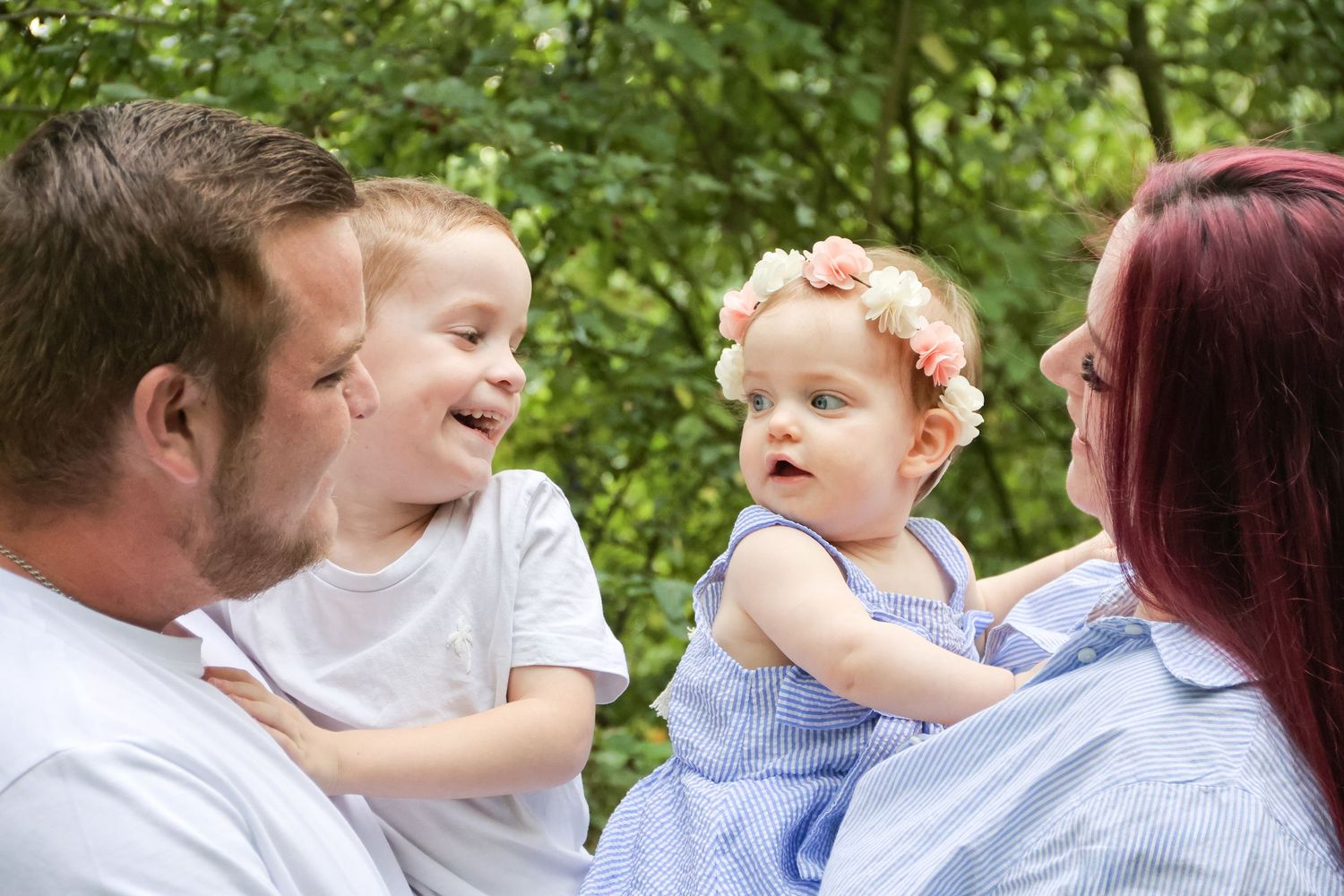 Happy family with two parents holding a laughing boy and baby girl wearing a floral headband outdoors.