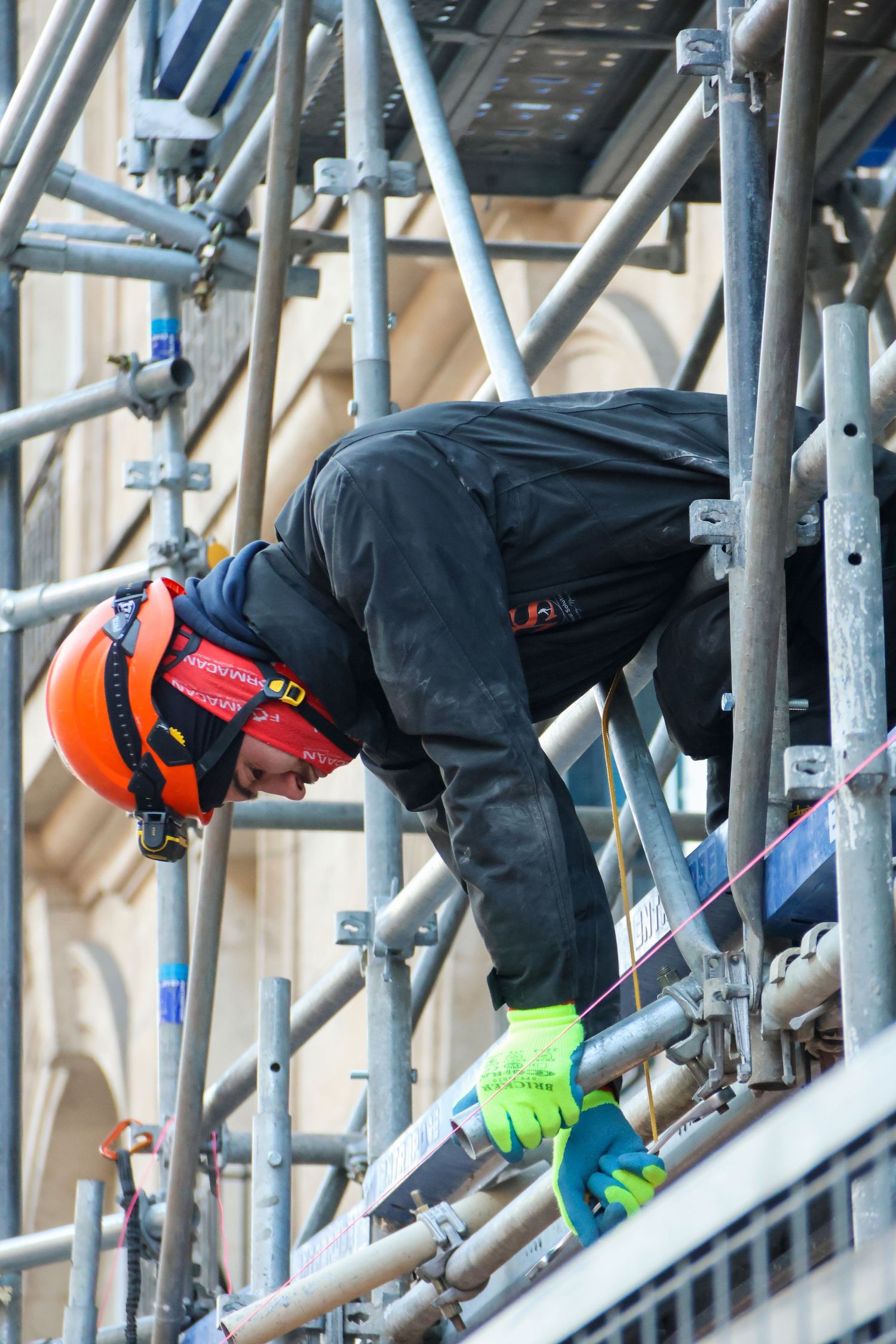 Construction worker in orange helmet and hi-vis gloves working on metal scaffolding at a building site.