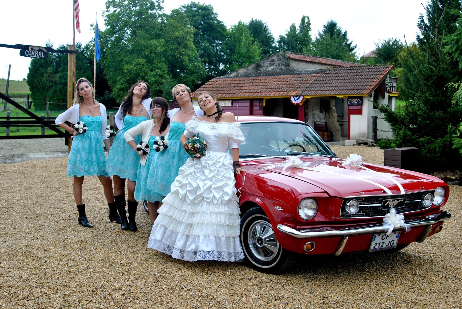 Bride in white ruffled gown and bridesmaids in teal dresses pose beside a decorated red vintage Mustang at a wedding.
