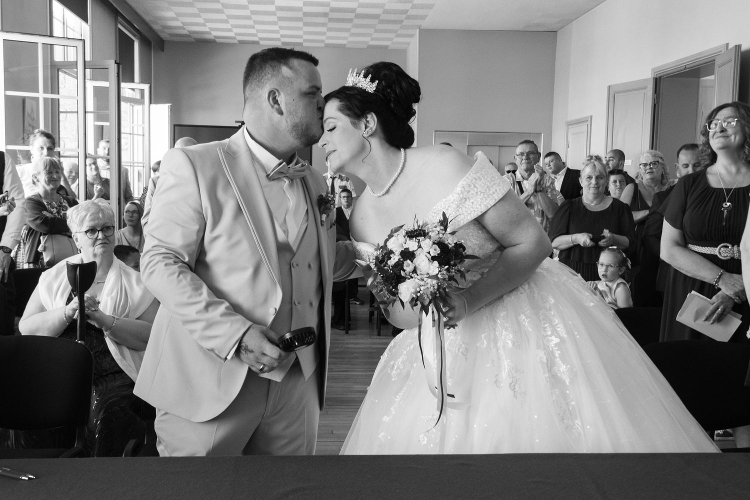 Black and white photo of a bride and groom sharing a kiss during their wedding ceremony with guests seated in background.