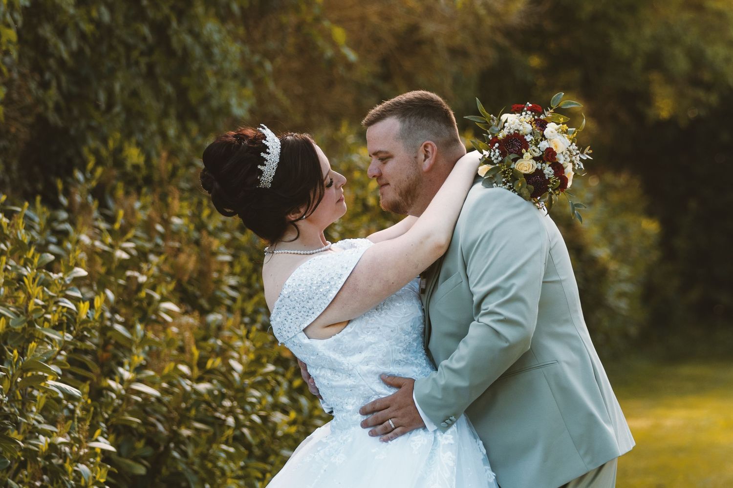 Bride and groom embrace outdoors, she wears a lace gown with tiara, he wears a sage suit, holding a floral bouquet.