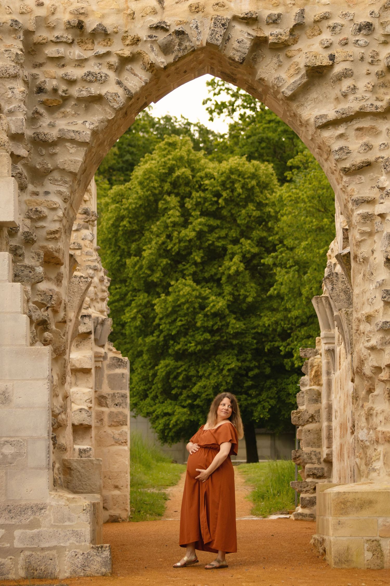Pregnant woman in rust dress standing under a stone arch ruin with lush green trees in background.