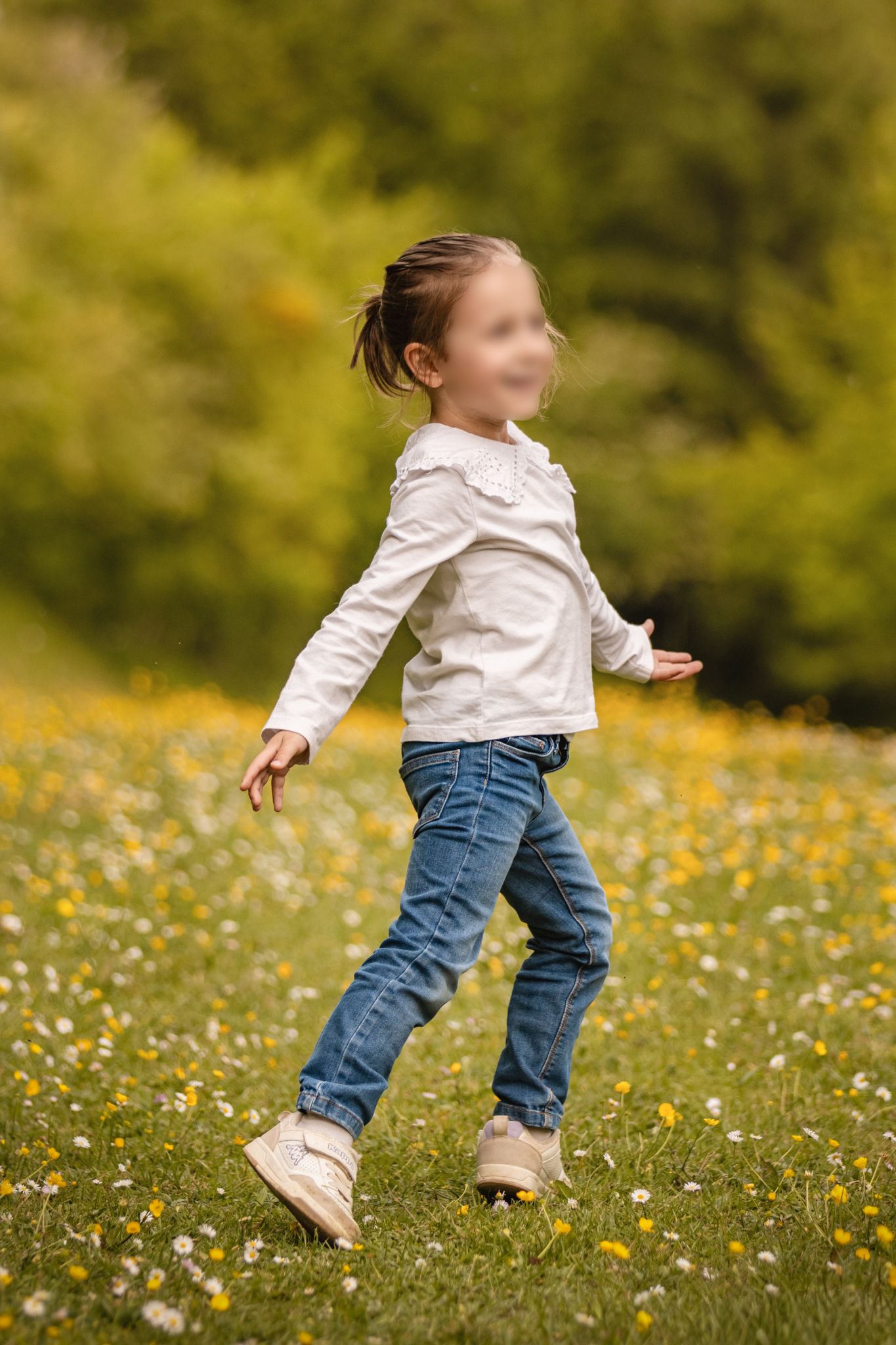 Young girl in white top and jeans running through a sunny meadow filled with wildflowers and green trees.