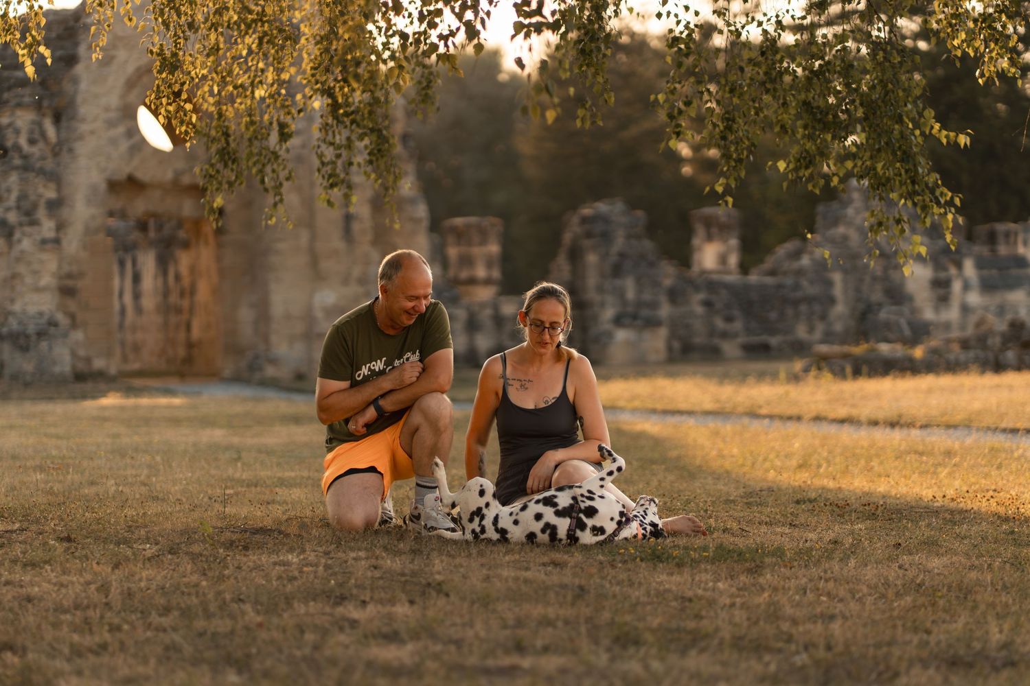 Couple sitting on grass with Dalmatian dog near ancient ruins during golden hour sunset.