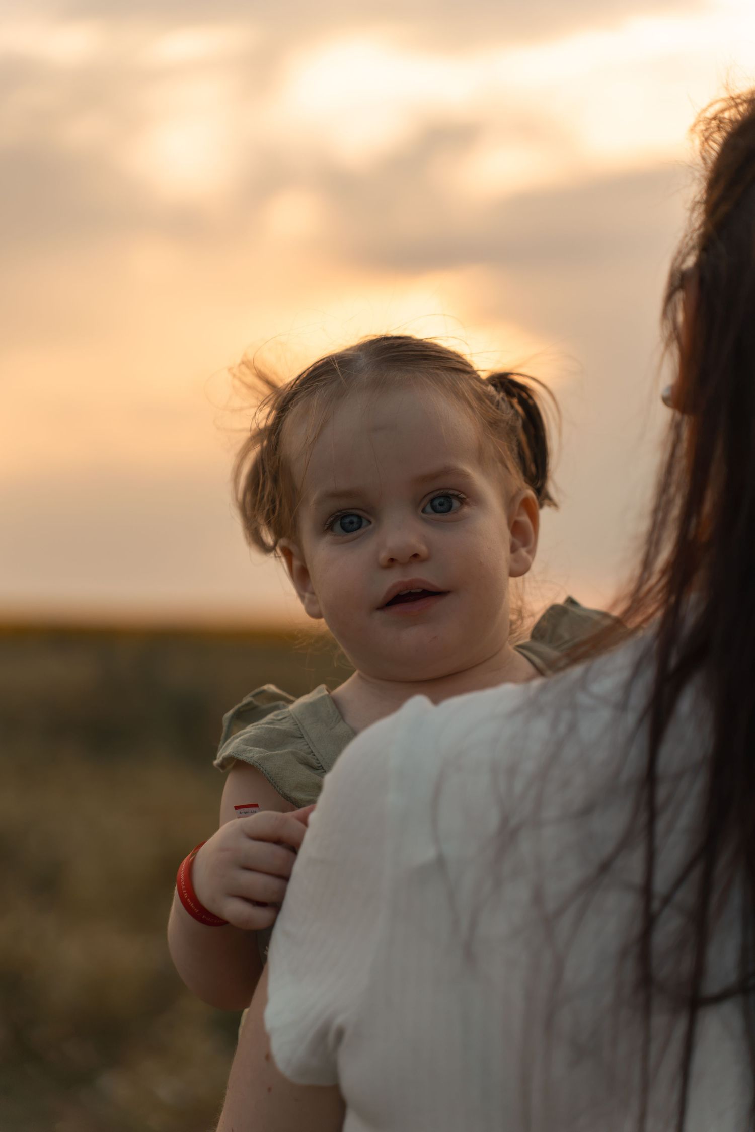 A toddler girl with blue eyes being held by her mother during a golden sunset outdoors.