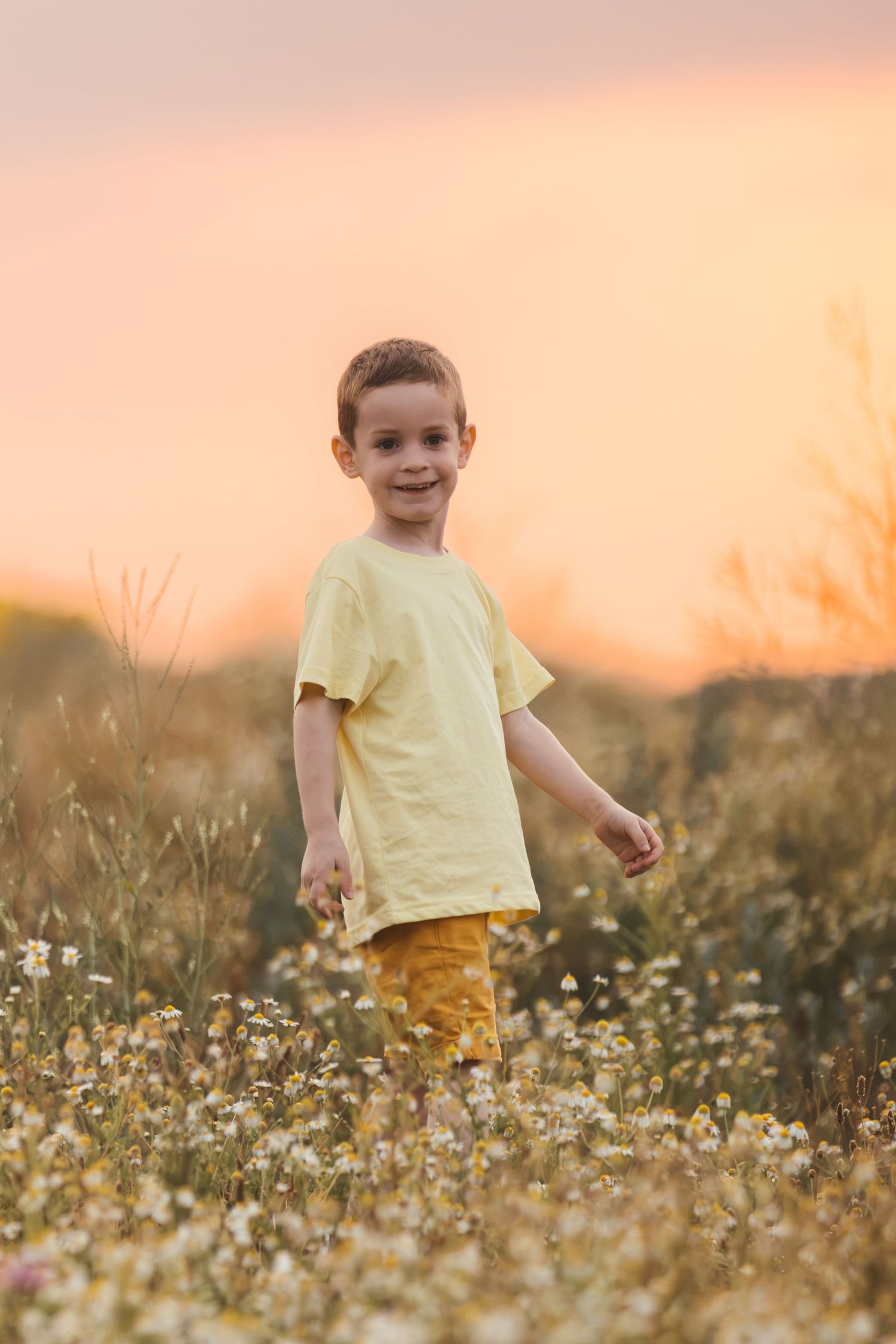 Young boy in yellow shirt standing in wildflower field at golden sunset, smiling at camera.