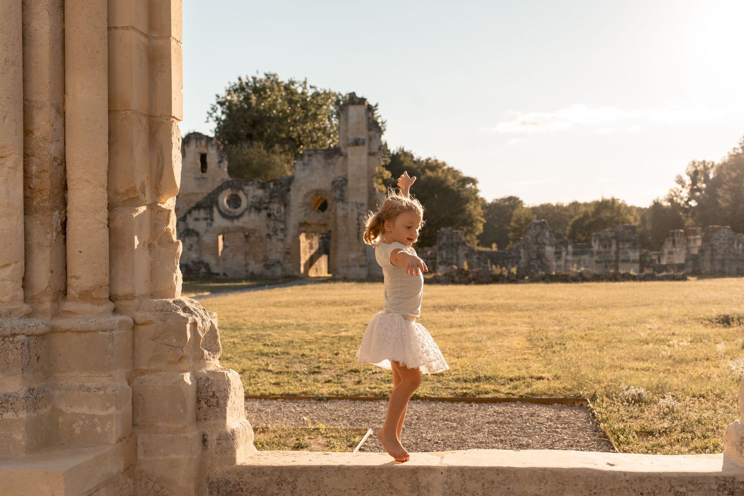 A barefoot little girl in a white dress dances near ancient stone ruins during golden hour.