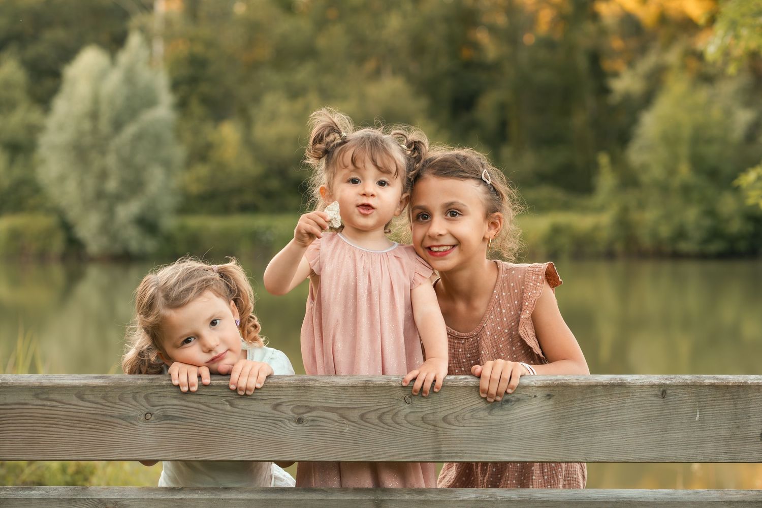 Three young sisters in pink dresses pose together at a wooden fence with a lake backdrop.