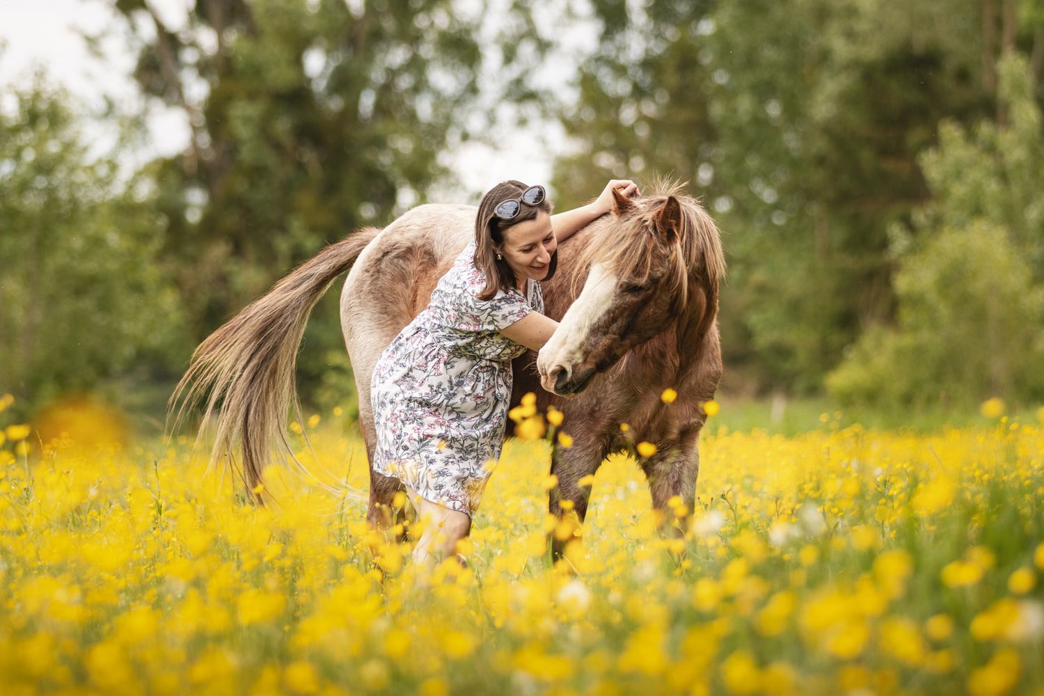 Woman in floral dress affectionately nuzzling a brown horse in a vibrant yellow wildflower meadow.