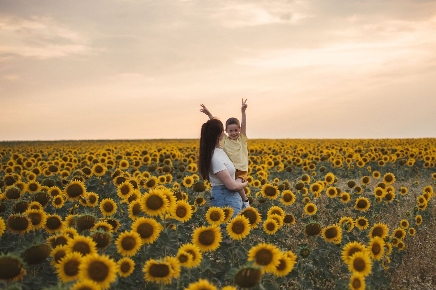 A mother holds her child in a vast sunflower field at sunset, both raising their hands joyfully.