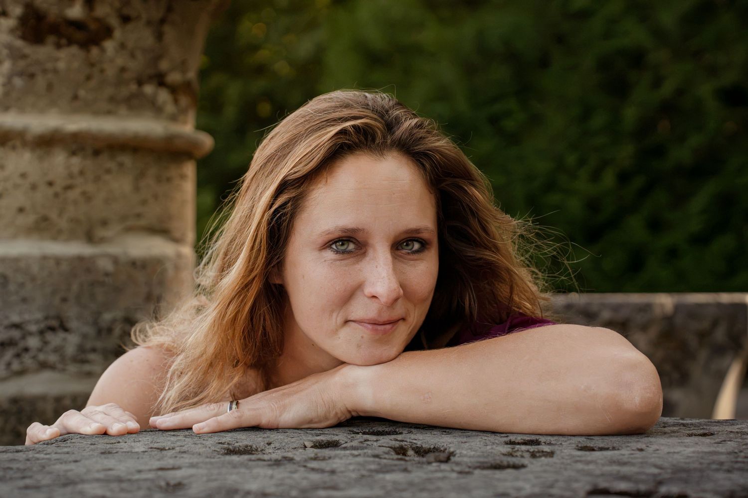 A woman with long wavy hair rests her chin on her arms atop a stone wall outdoors.