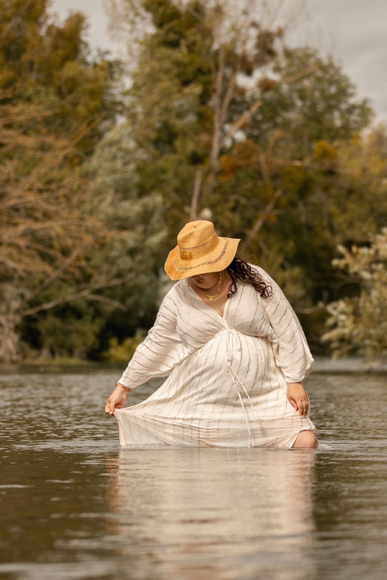 Pregnant woman in white dress and straw hat kneeling in river water, looking down at her dress.