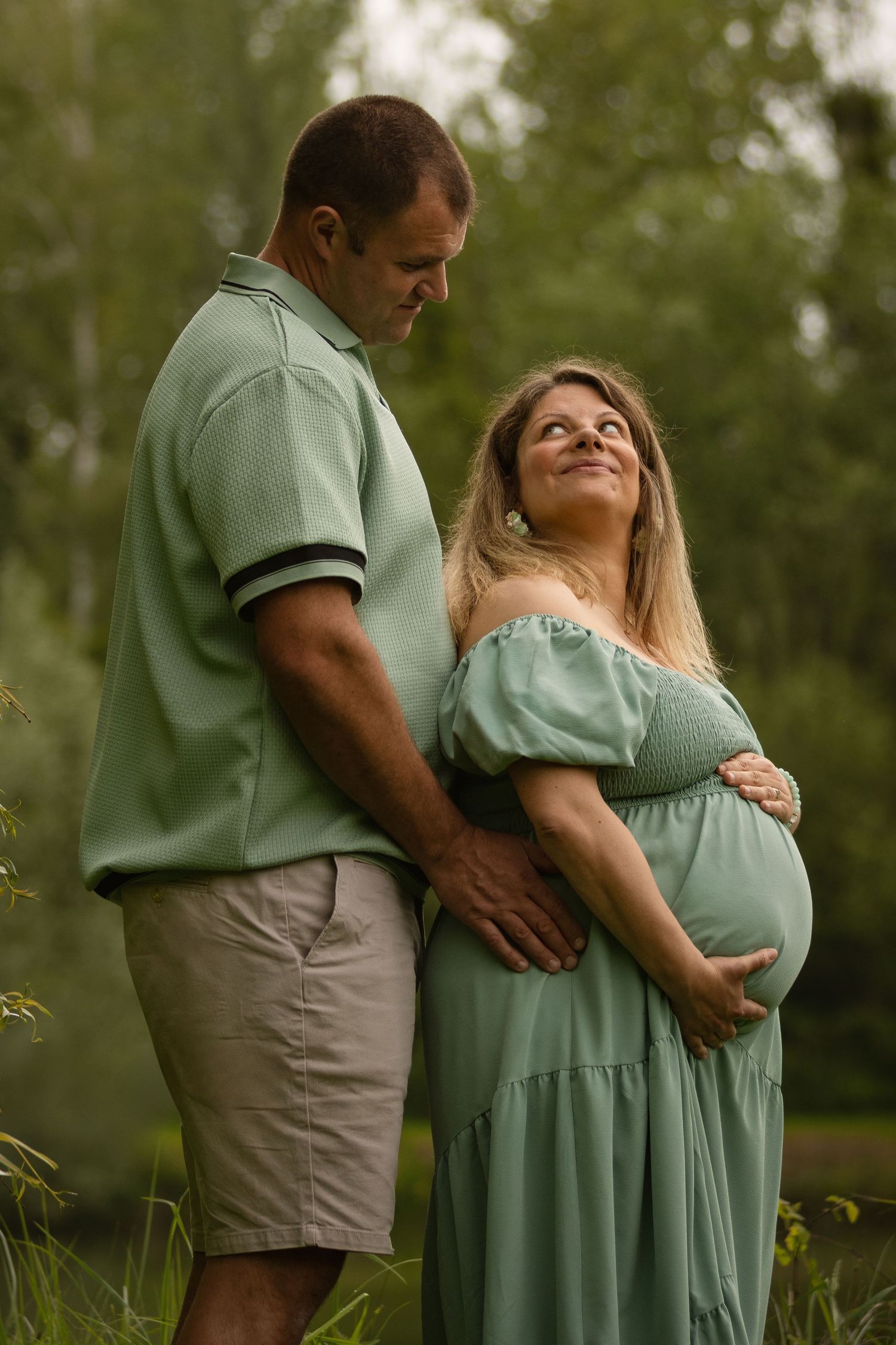 A couple in matching green outfits pose outdoors, the man embracing his pregnant partner in a lush garden setting.