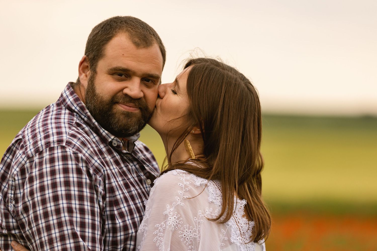 Woman kissing bearded man on the cheek while he smiles in a sunny outdoor field setting.