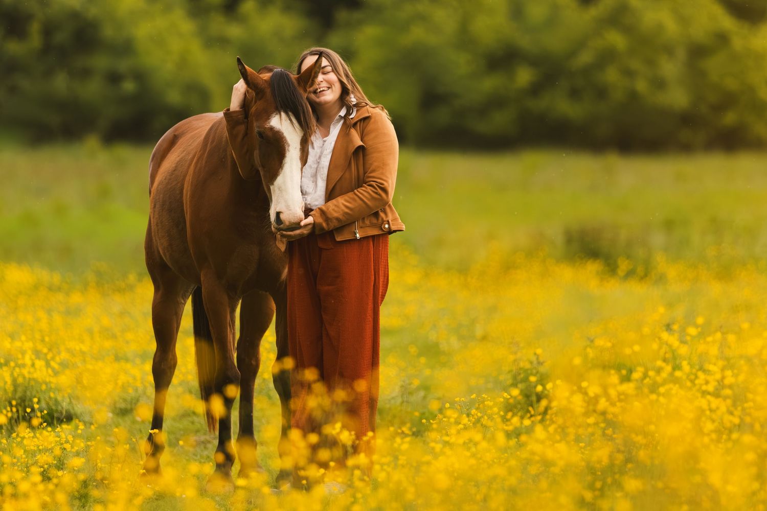 Woman in rust dress smiling beside chestnut horse in vibrant yellow wildflower field on sunny day.