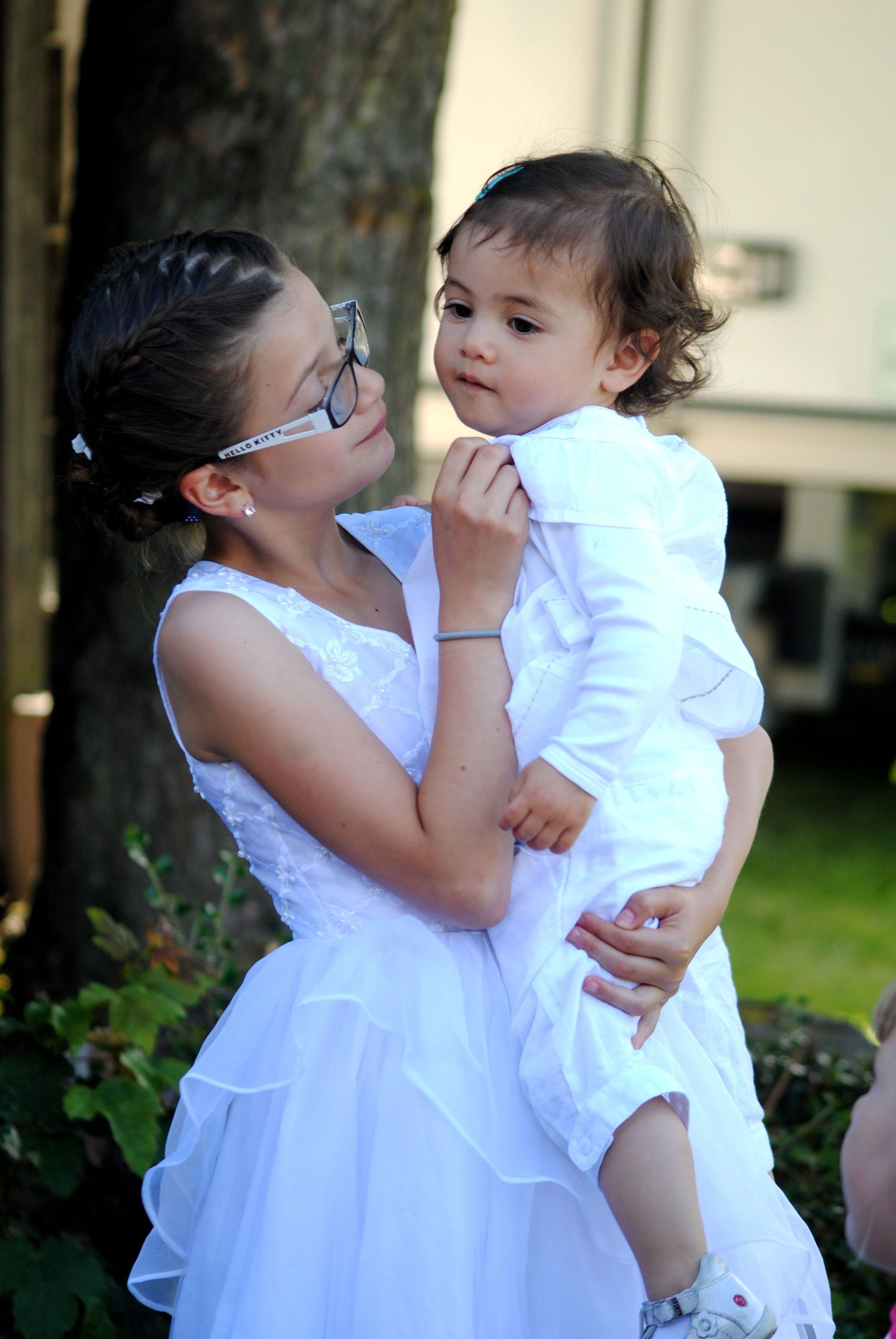A young girl in a white dress and glasses tenderly holds a baby dressed in white at an outdoor event.
