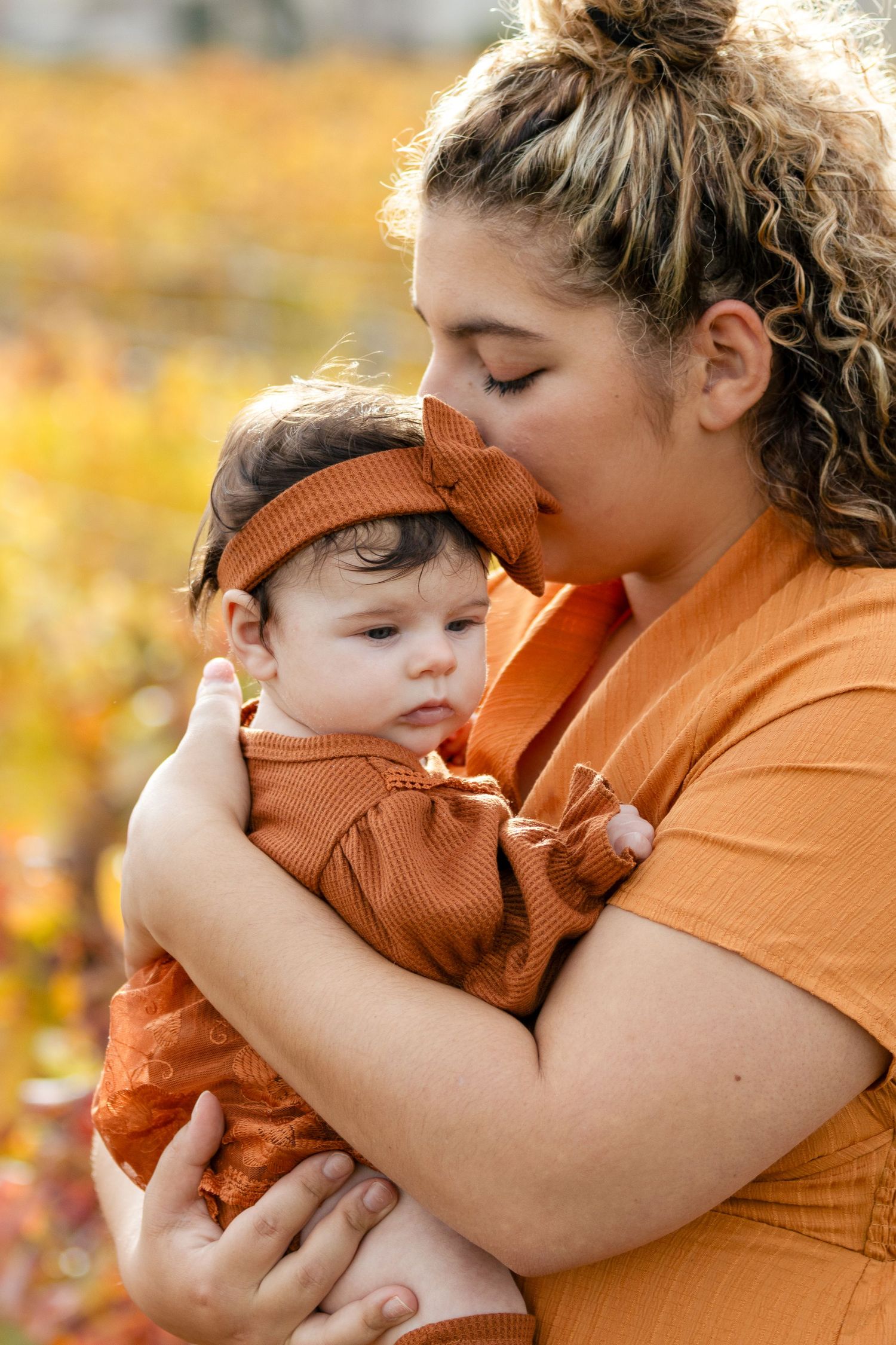 Mother in orange dress kissing her newborn baby dressed in rust-colored outfit outdoors.