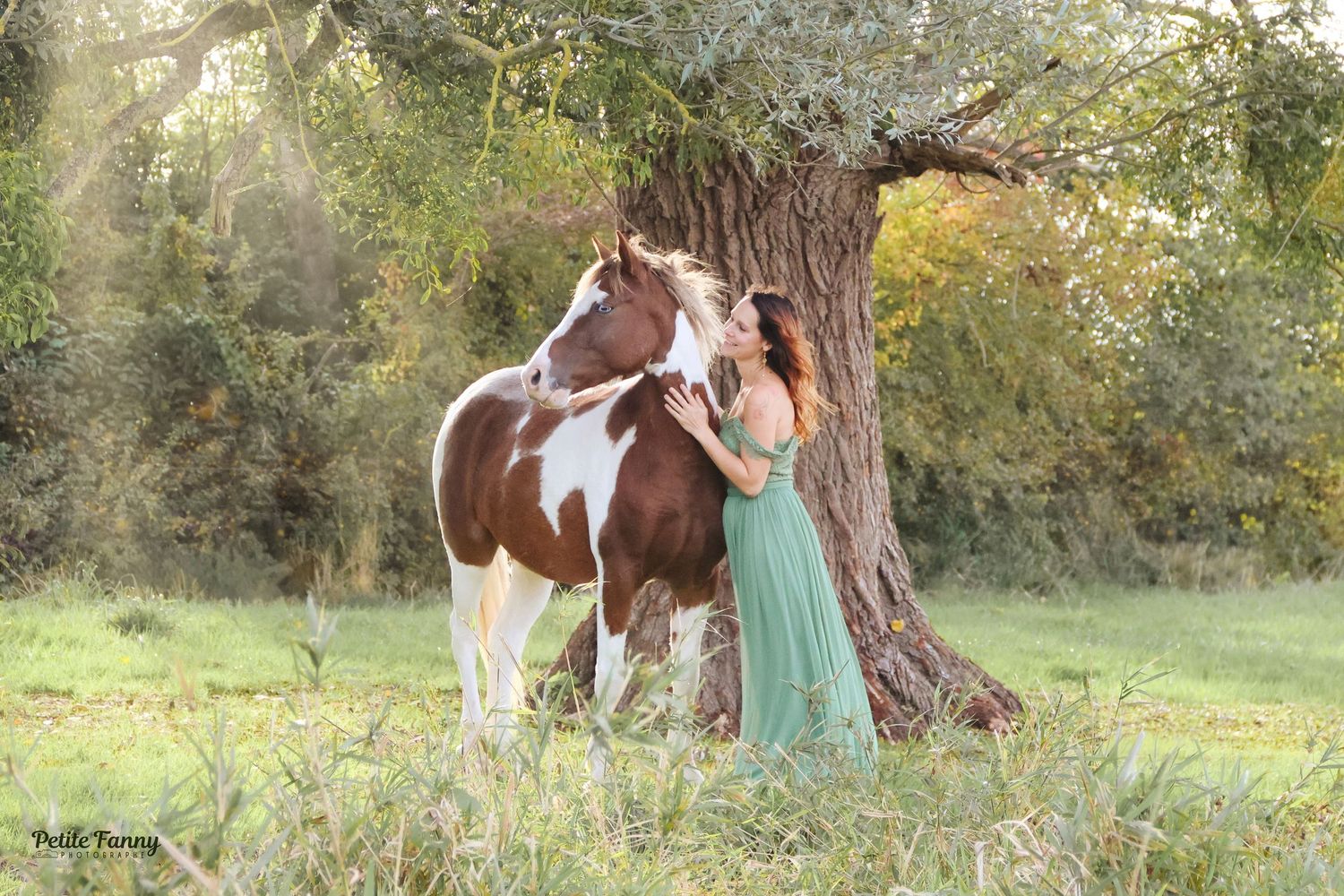 Woman in green dress standing beside a brown and white paint horse under a tree.