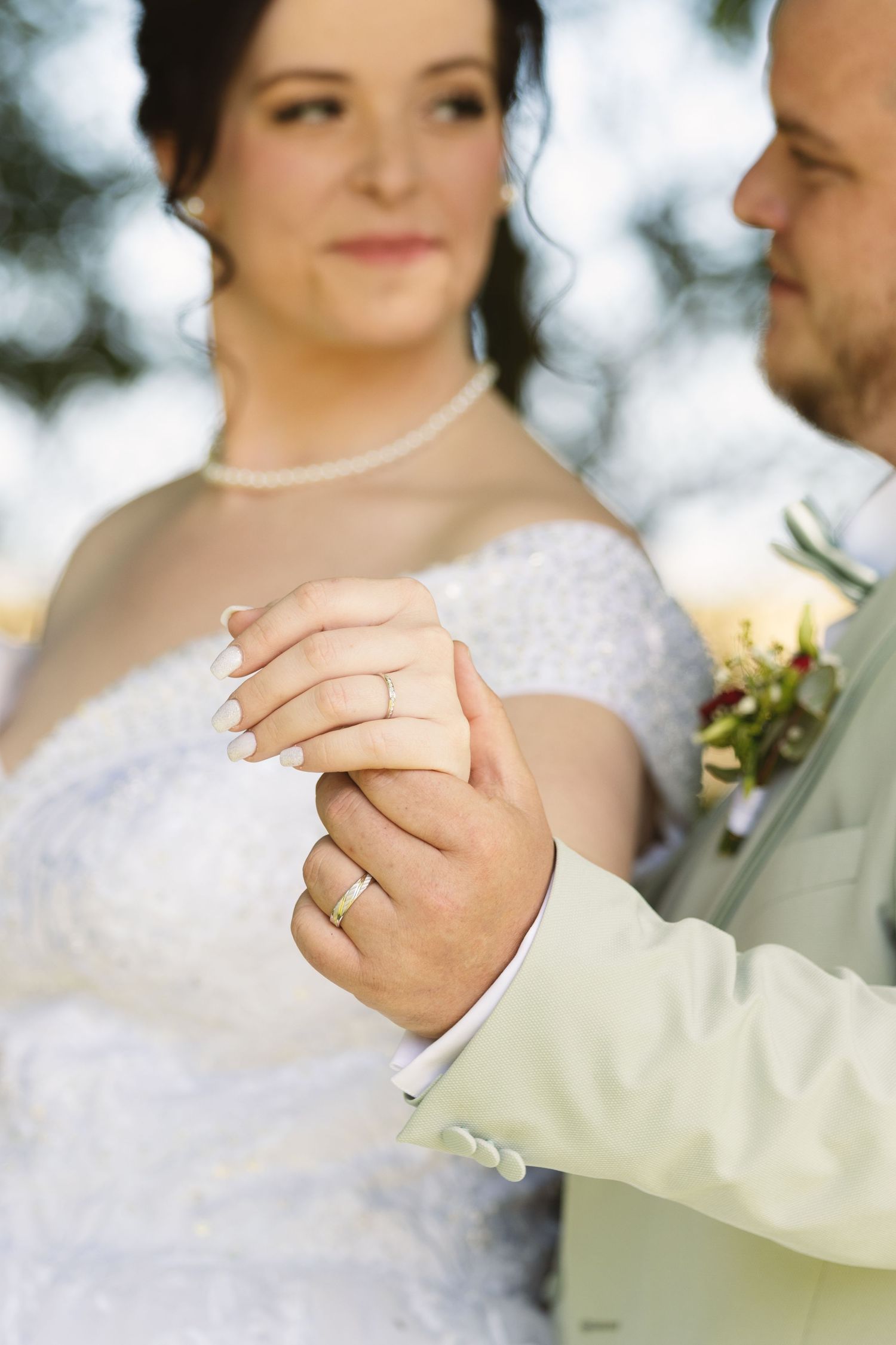 Bride and groom holding hands showing wedding rings, bride in white lace dress smiling softly outdoors.