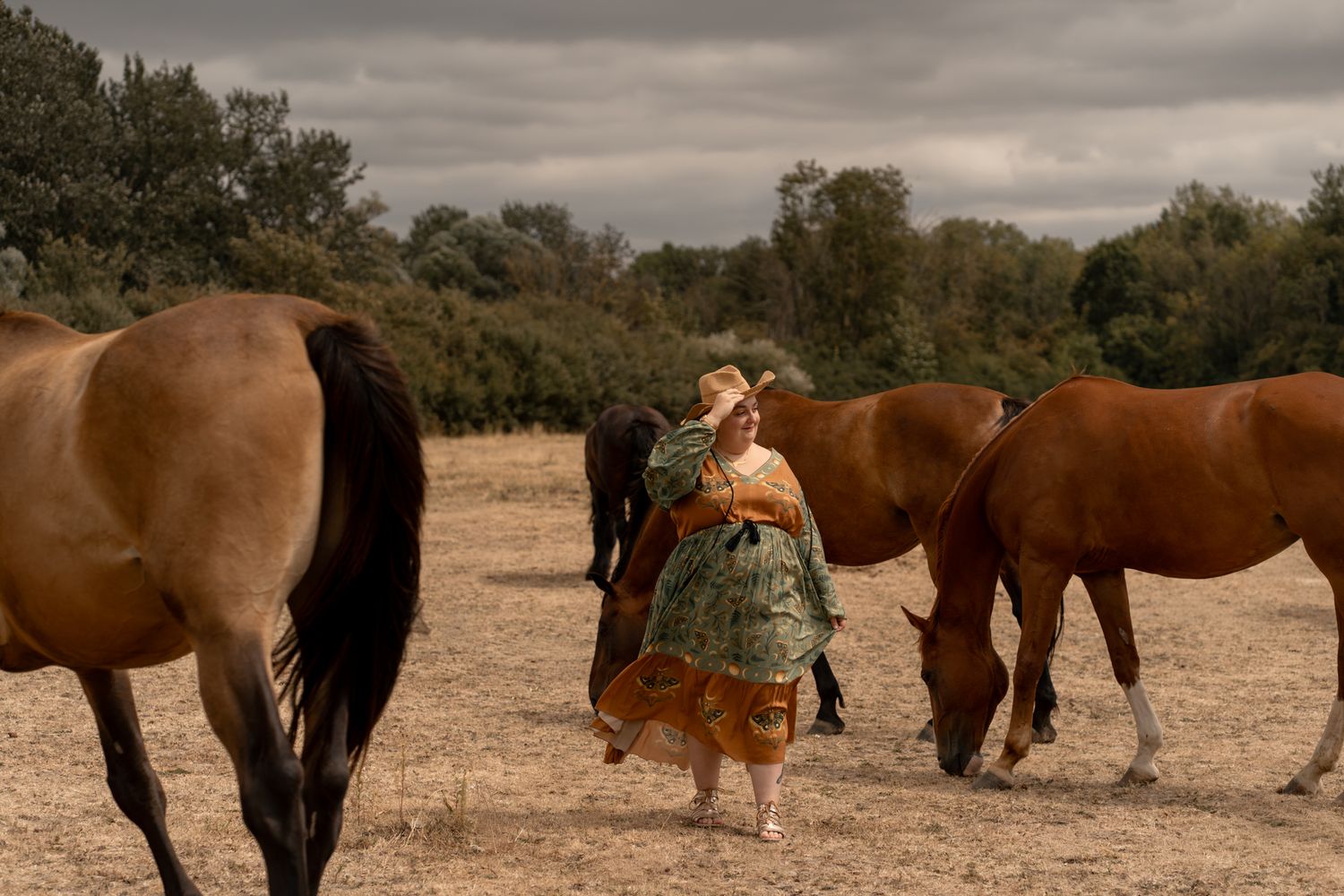 Woman in colorful dress walking among grazing chestnut horses in an open field under cloudy skies.