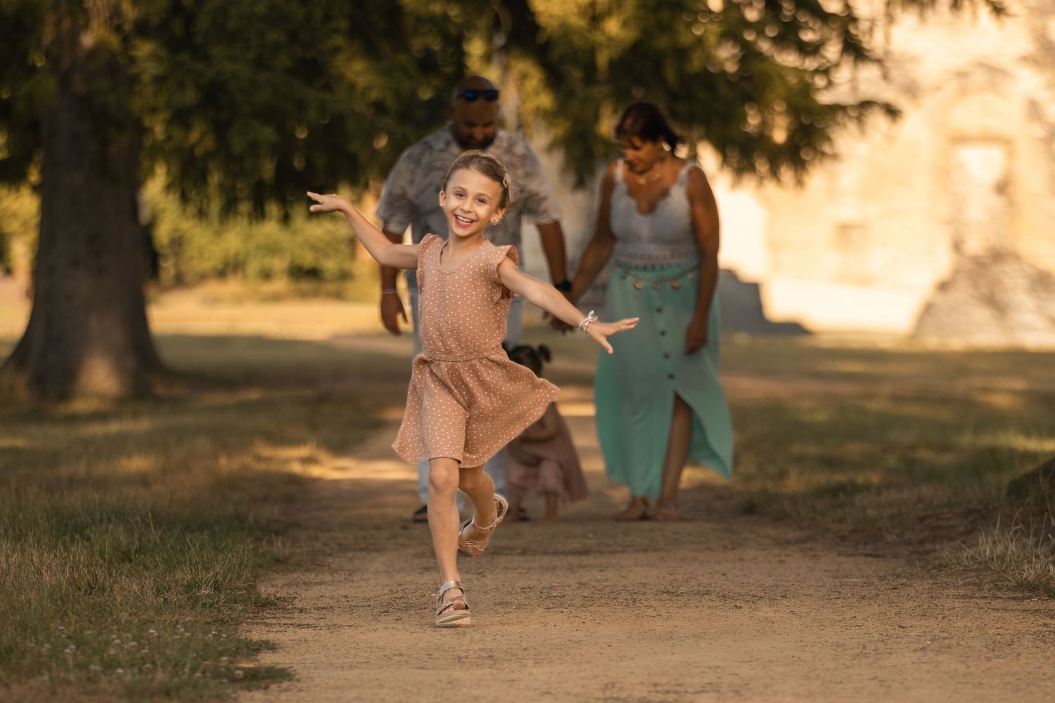A joyful girl in a pink dress runs ahead of her parents on a sunny park path surrounded by trees.