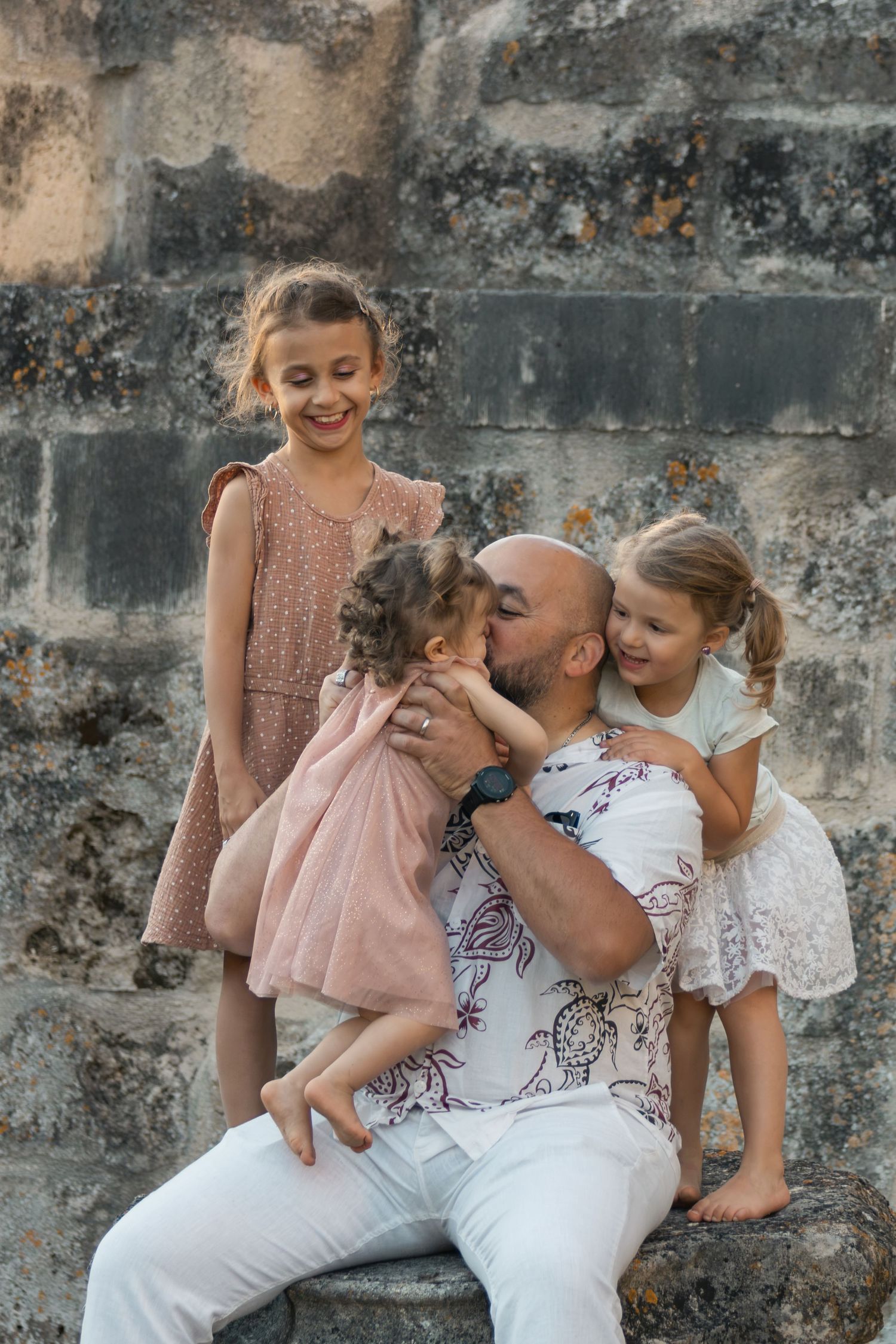 Bald father kisses toddler while two daughters smile during family photo session by stone wall.