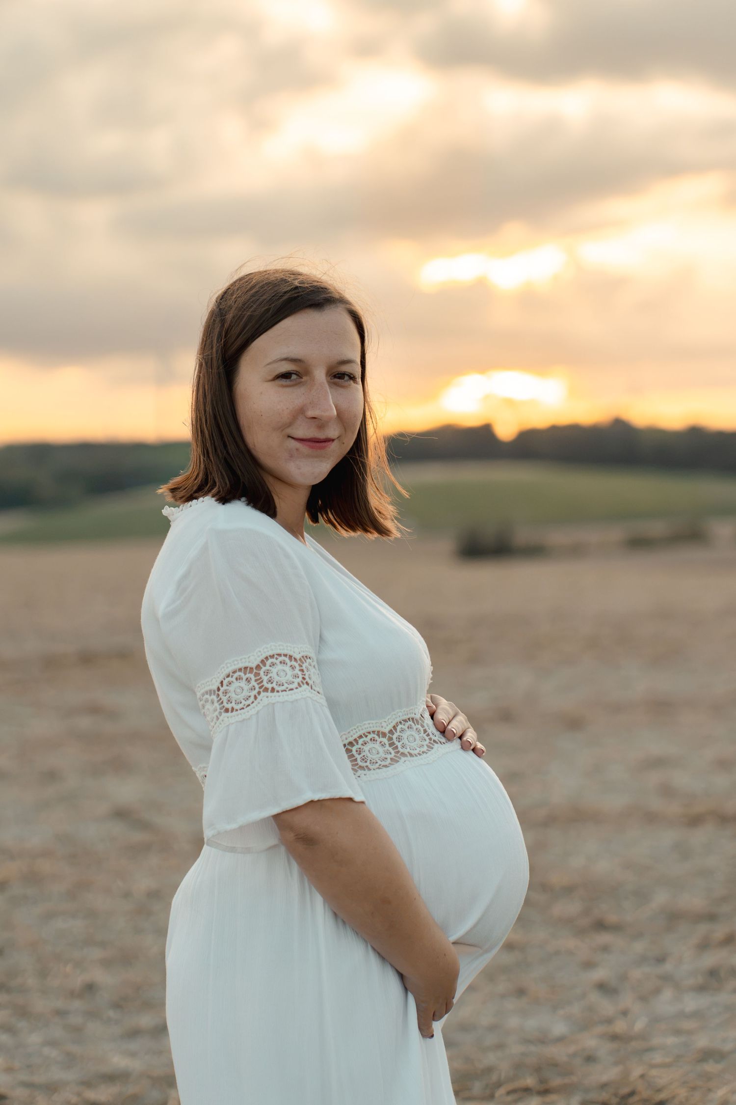 Pregnant woman in white dress holding baby bump at golden sunset in a field.