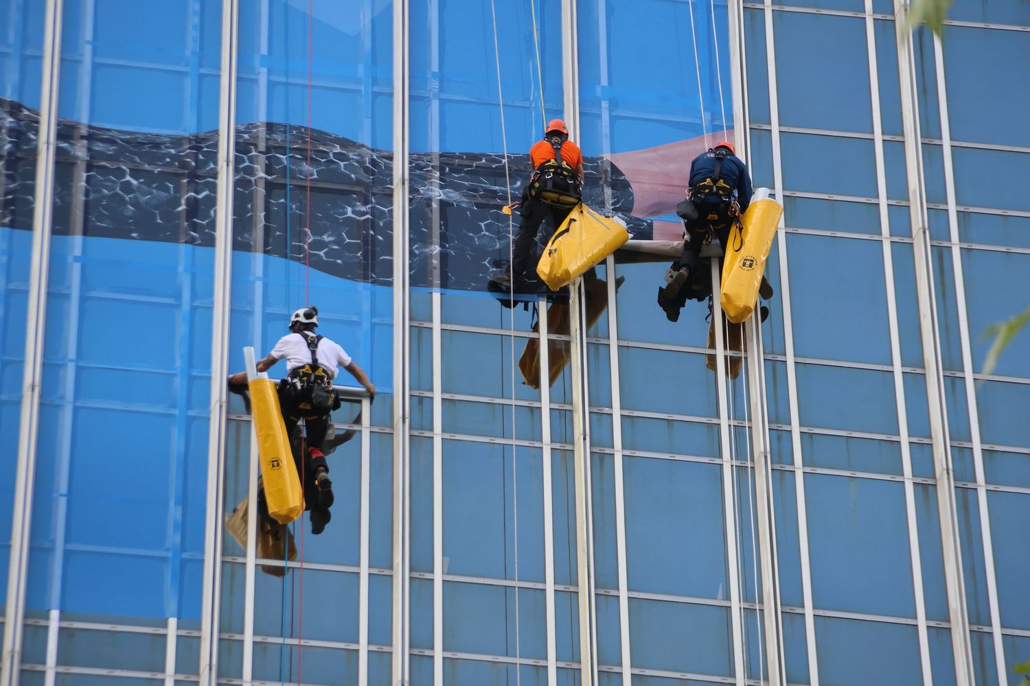 Window washers suspended on scaffolding clean the glass facade of a tall blue mirrored skyscraper.