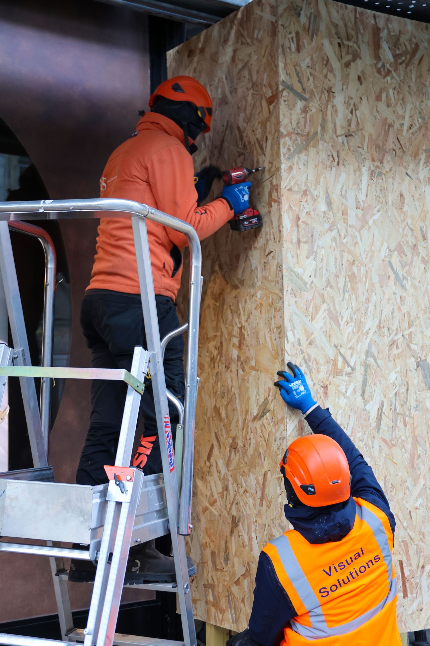 Two Visual Solutions workers in orange helmets installing OSB board on a wall, one on a ladder using a power drill.