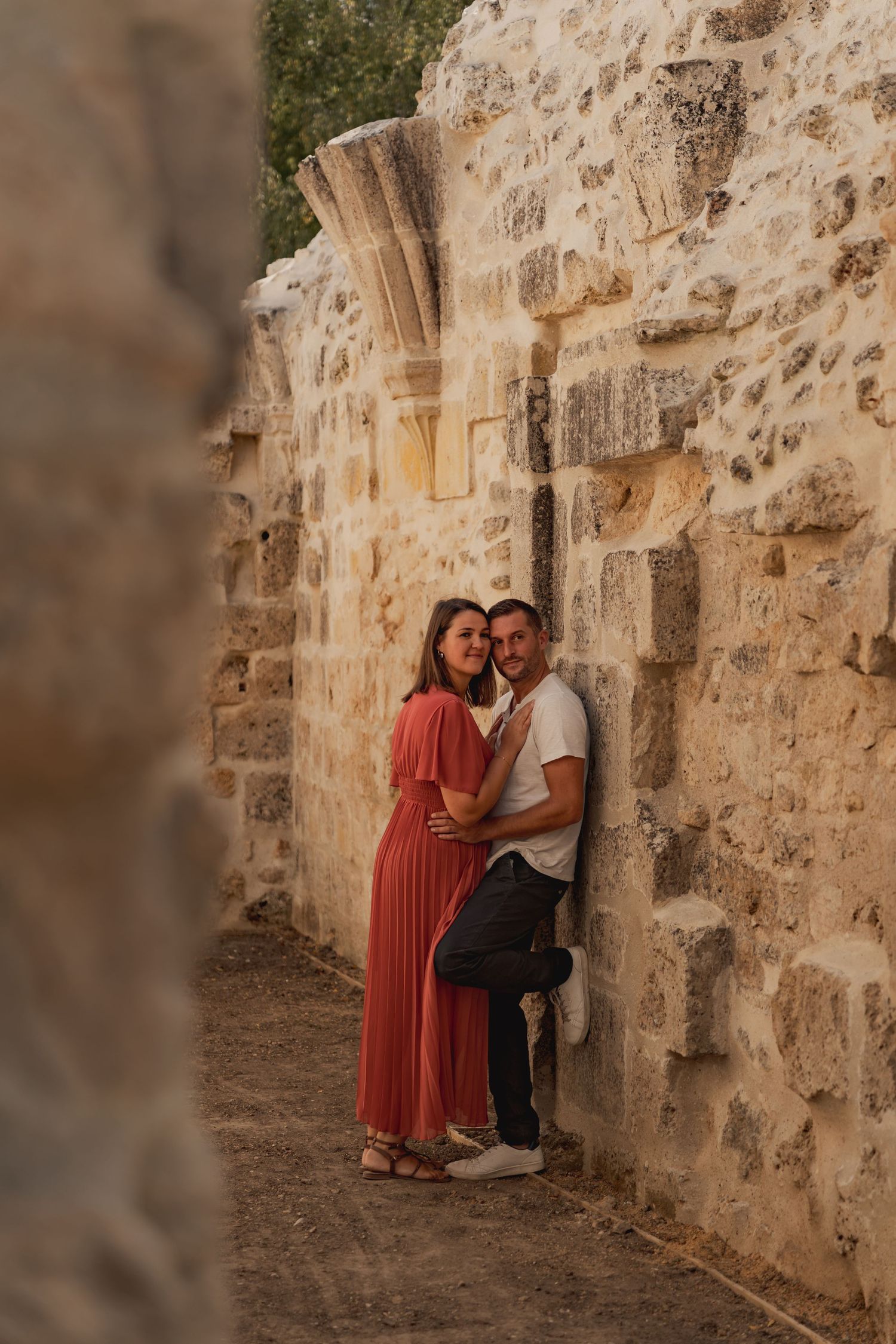 Couple embracing against ancient stone wall ruins, woman in red dress, man in white shirt, warm golden light.